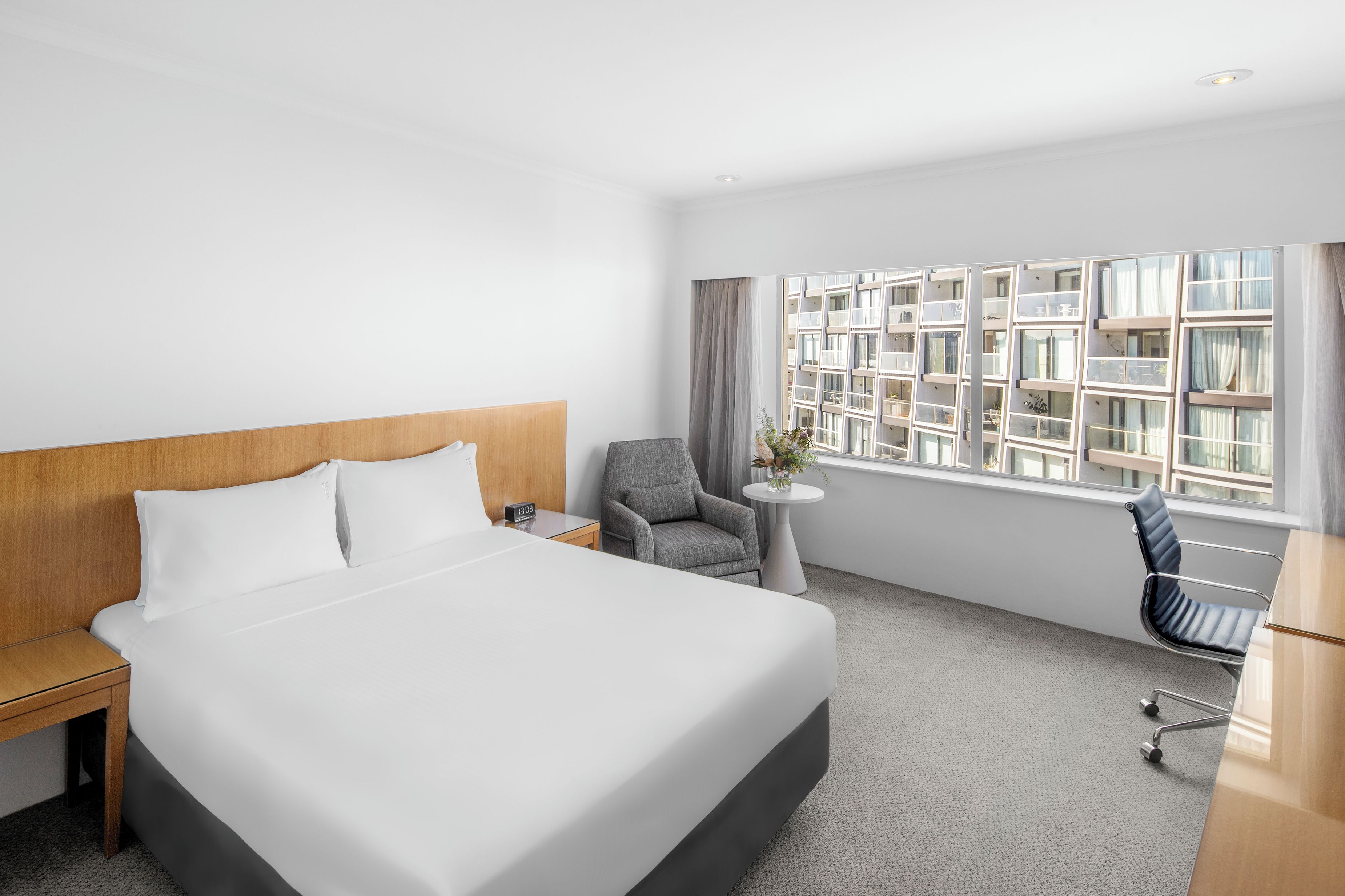 Habitación de hotel con una cama grande, un escritorio, una silla, una mesita con un florero y una ventana con vistas a edificios.