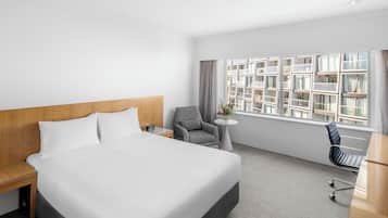 Habitación de hotel con una cama grande, un escritorio, una silla, una mesita con un florero y una ventana con vistas a edificios.