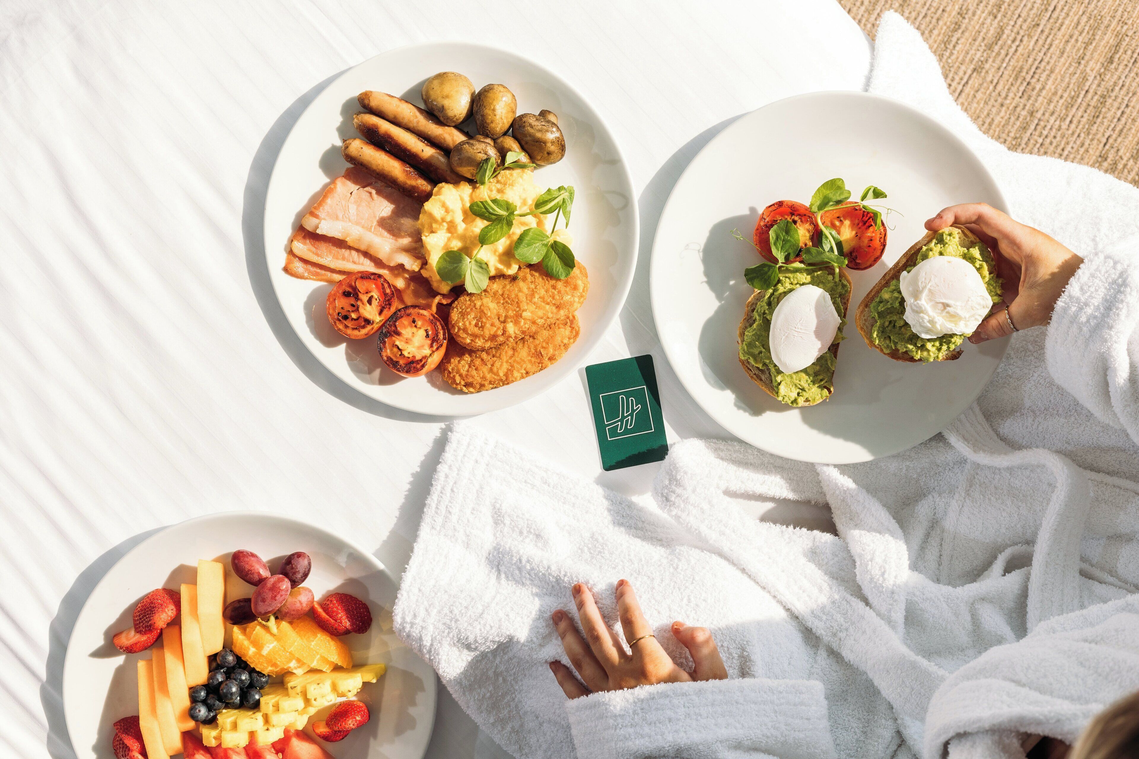 A person in a white bathrobe lying on a white towel with plates of food, including toast with avocado and a fruit platter.