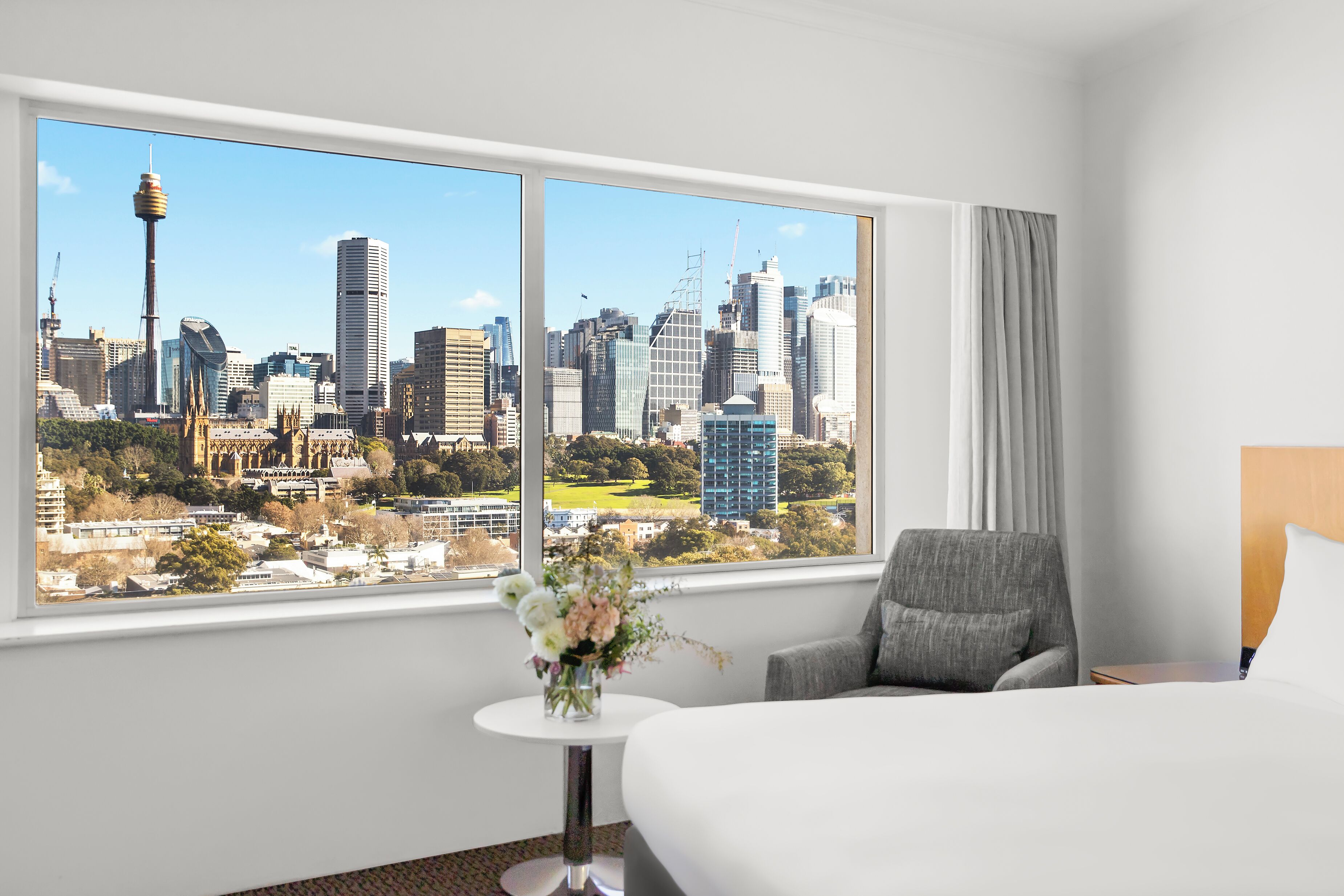 Quarto de hotel com uma grande janela que oferece vista para a cidade, uma cama, uma cadeira e uma pequena mesa com um vaso de flores.