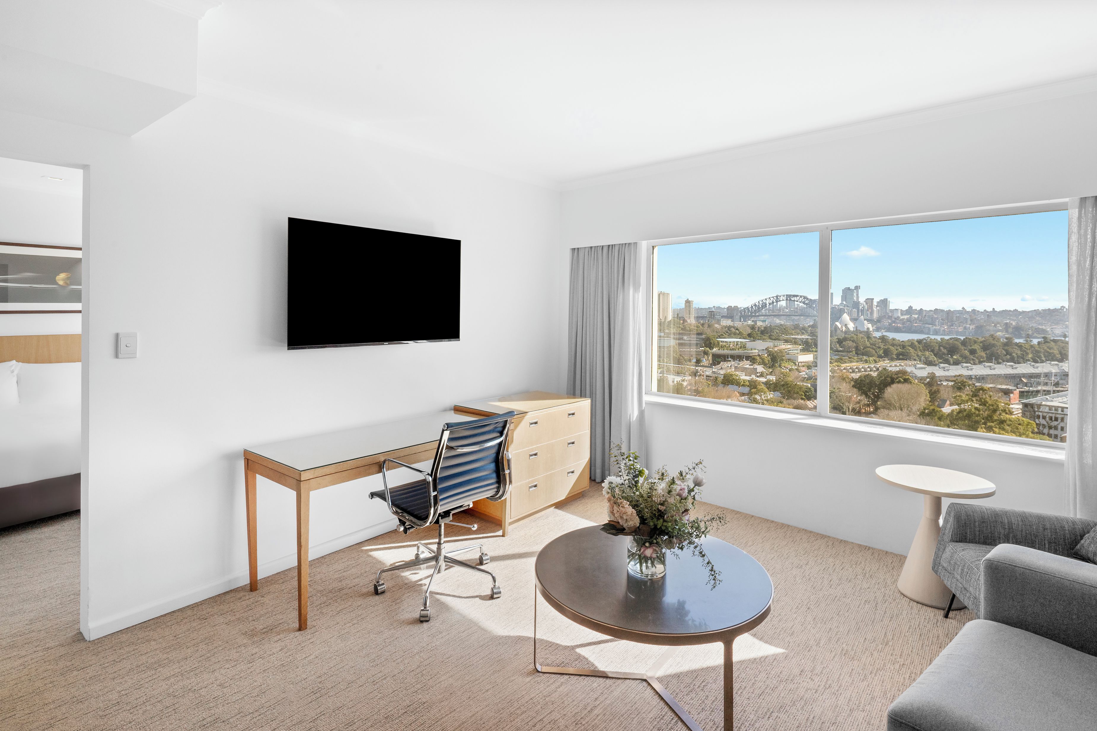 A modern hotel room with a large window, a flat-screen TV, a desk, a chair, a dresser, a round coffee table, and a sofa.