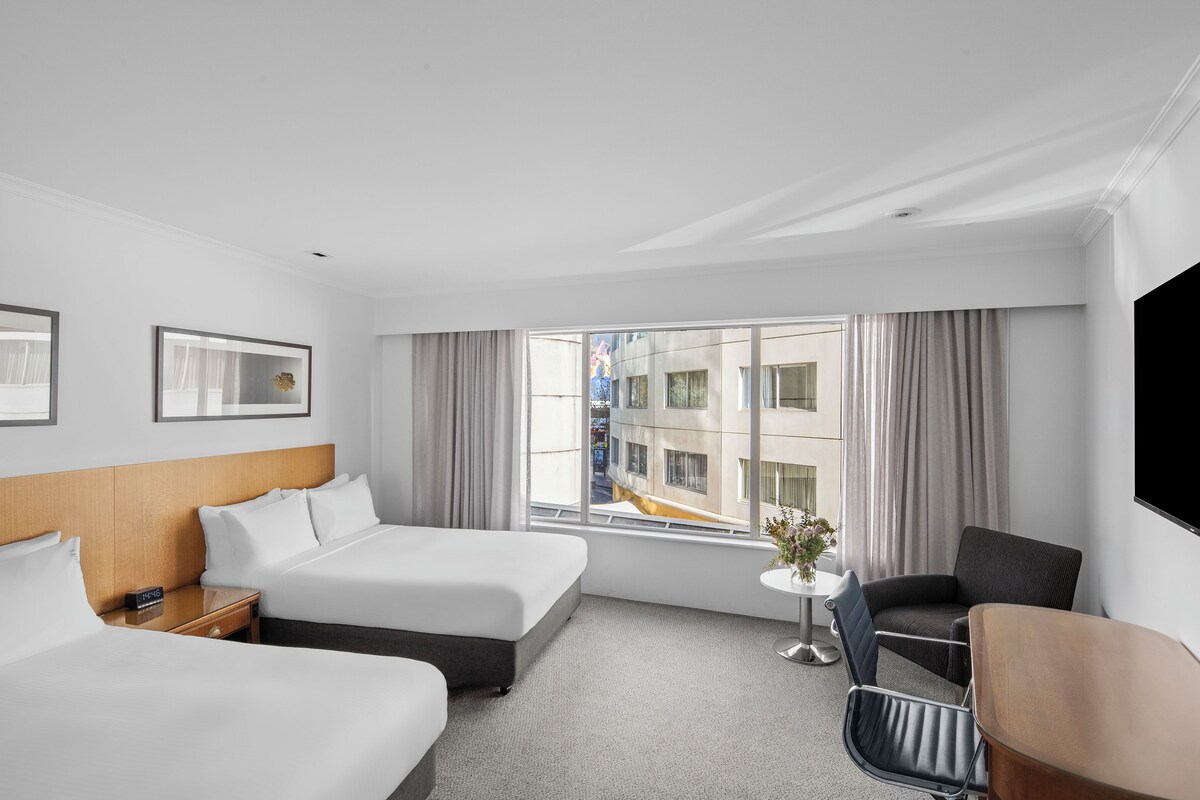 A hotel room with two beds, a desk, a chair, a TV, and a window with curtains.