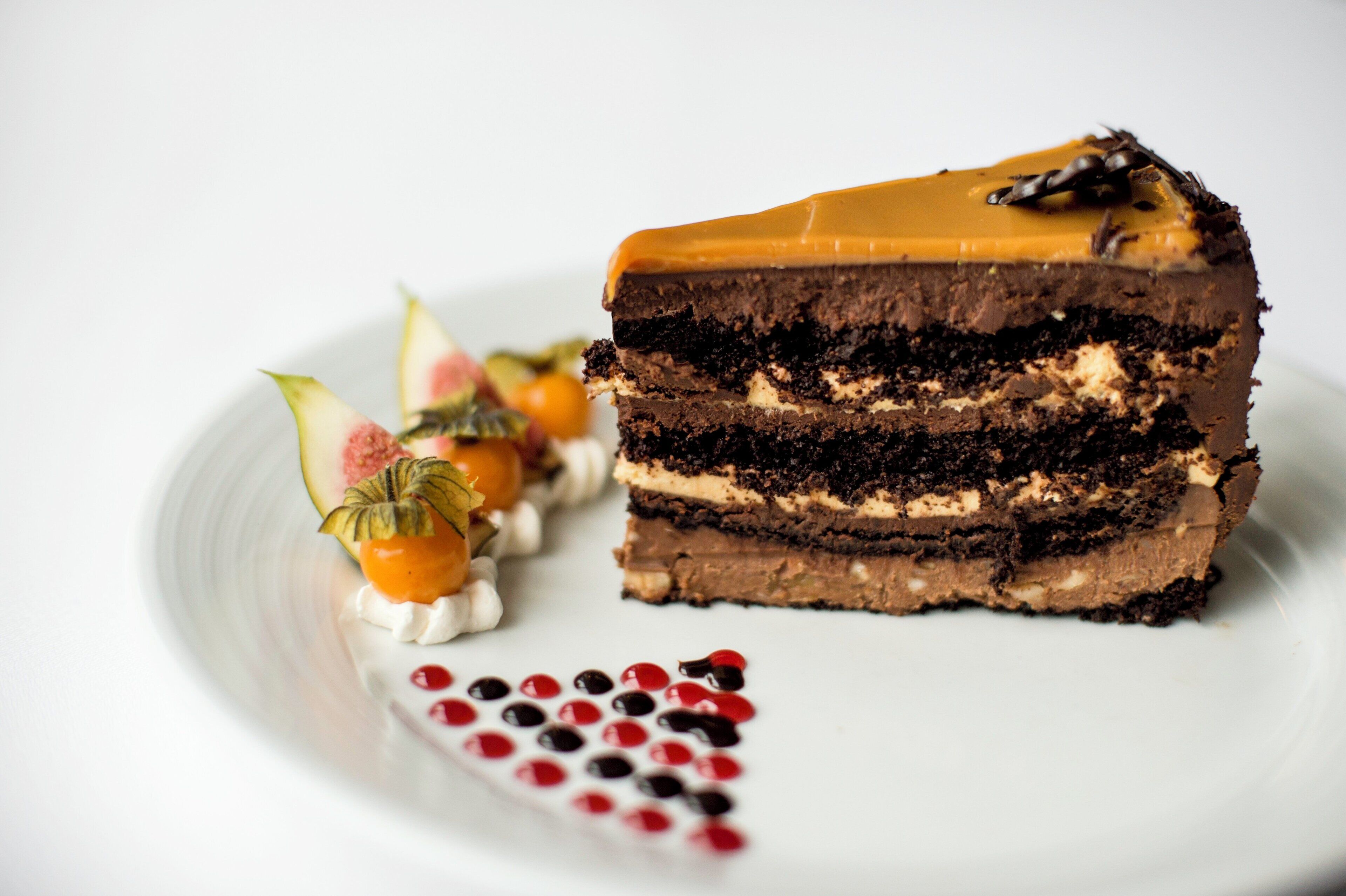 A slice of layered chocolate cake with a caramel topping, accompanied by decorative fruit and berries on a white plate.