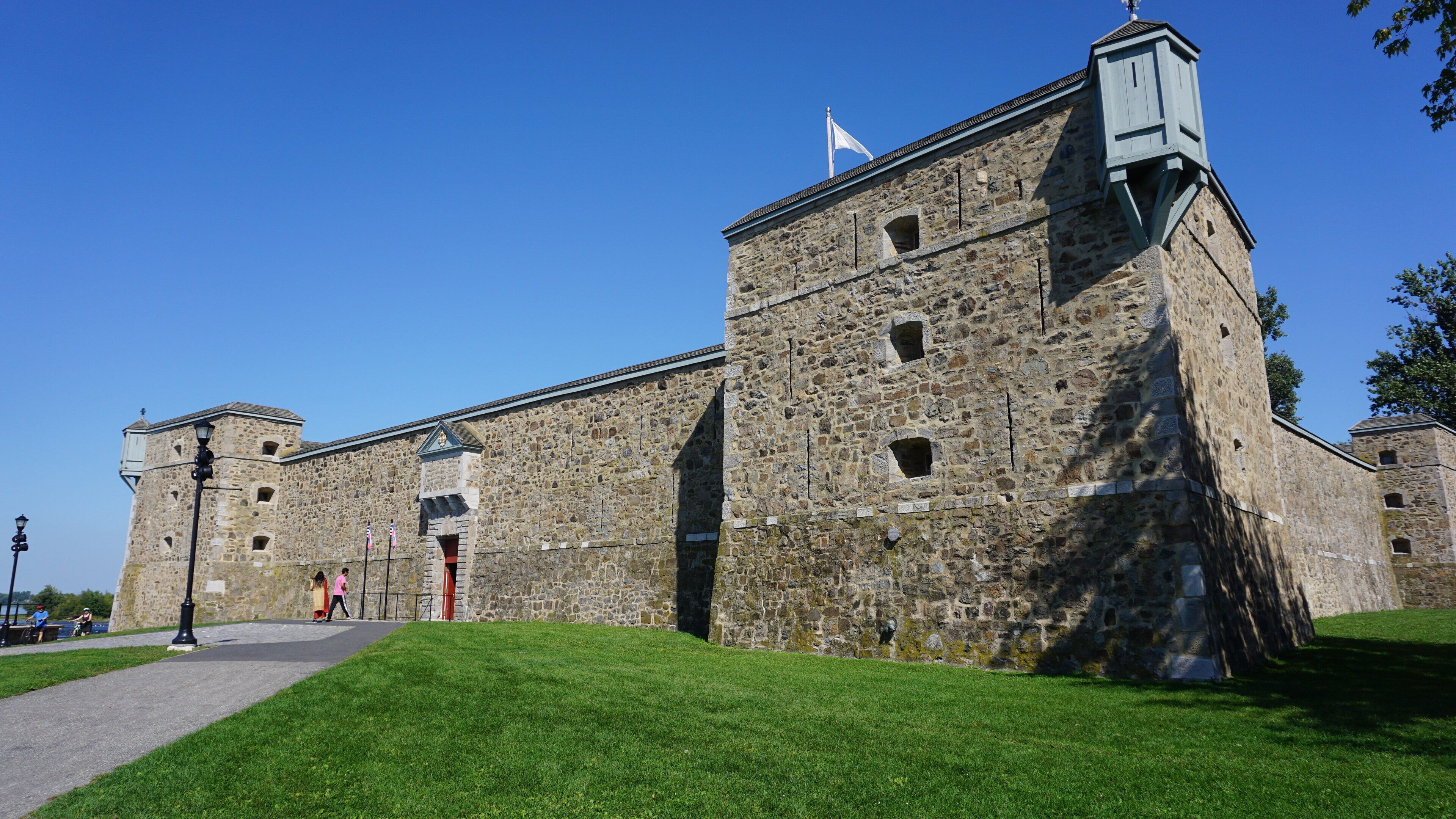 A historic stone building with a flag on top, a flagpole, and a paved walkway.