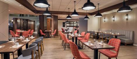 A modern restaurant interior with multiple dining tables and chairs, pendant lighting, and a wooden floor.