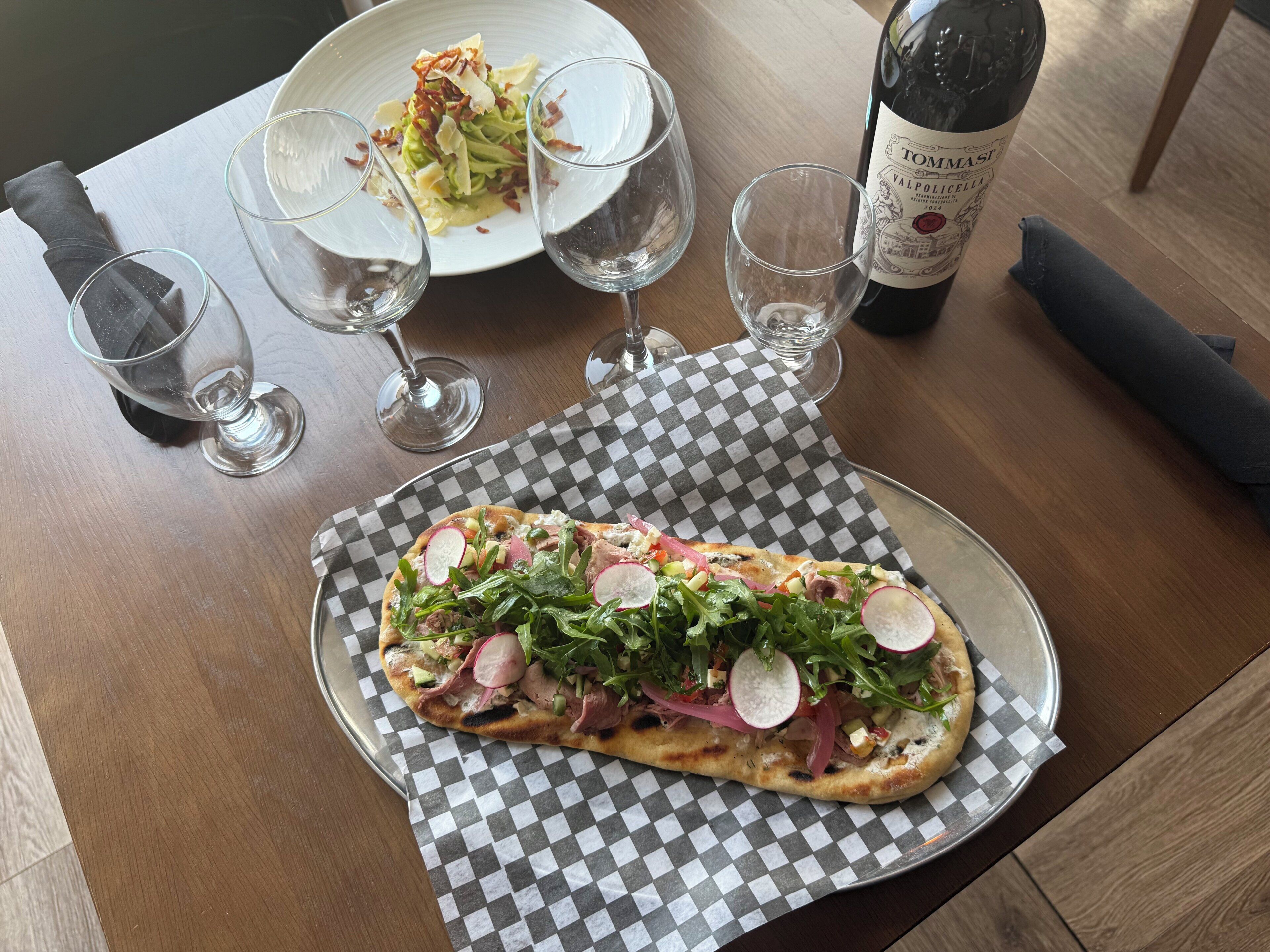 A pizza on a checkered paper, a bottle of wine, and empty wine glasses on a wooden table.