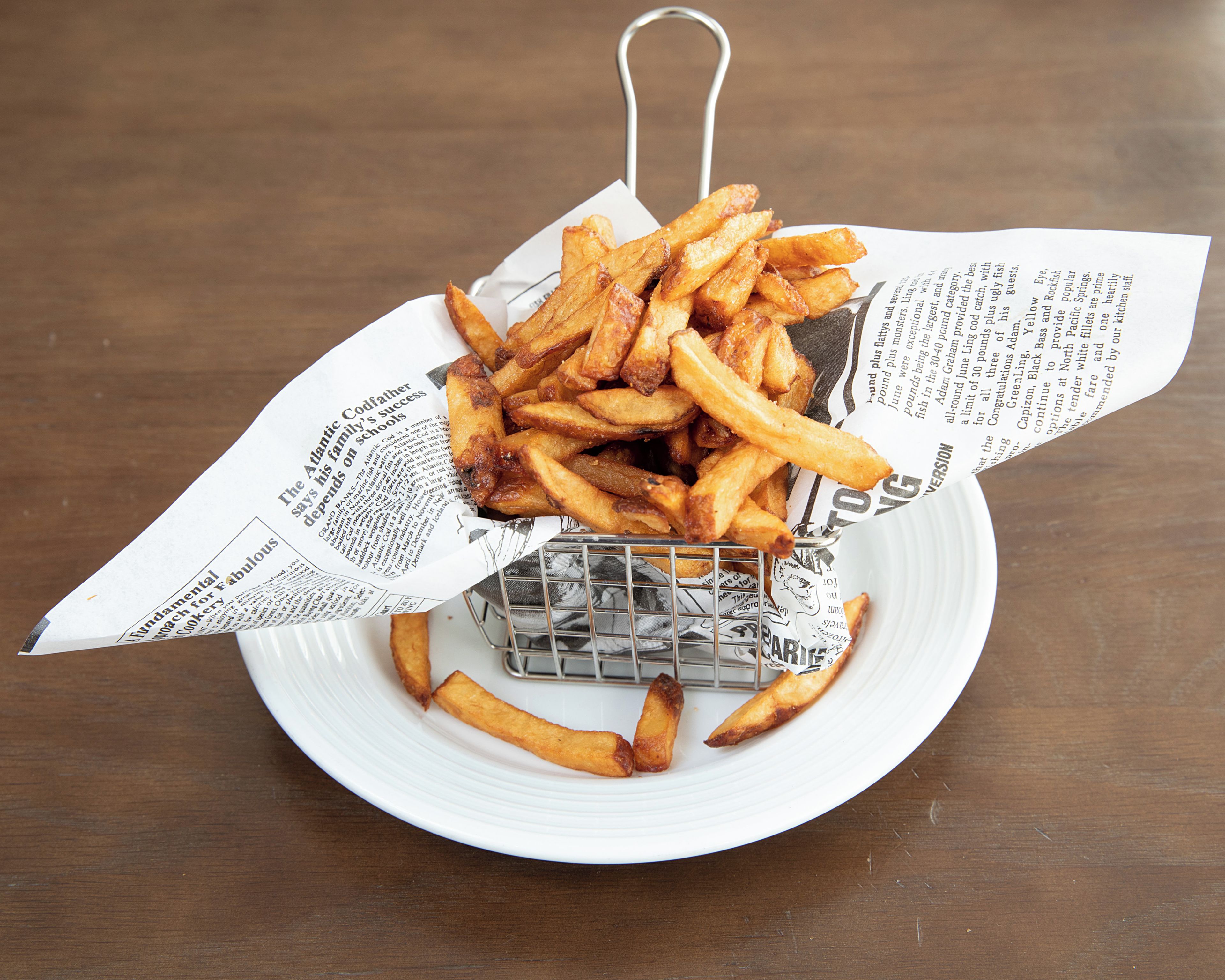A plate with a basket of golden-brown fries, a metal strainer, and a newspaper underneath.