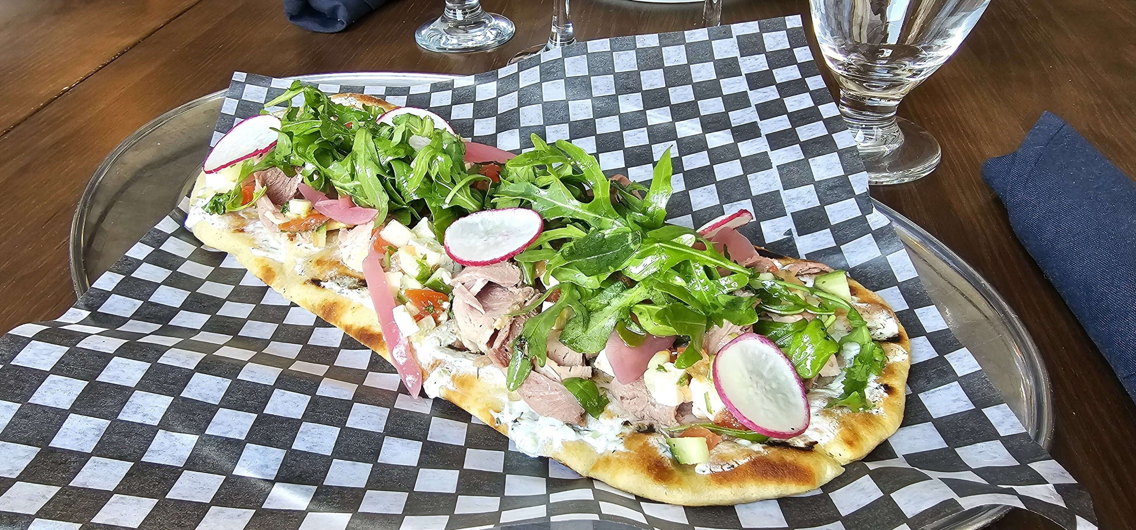 A checkered tablecloth with a glass plate containing a sandwich with greens, radishes, and meat.