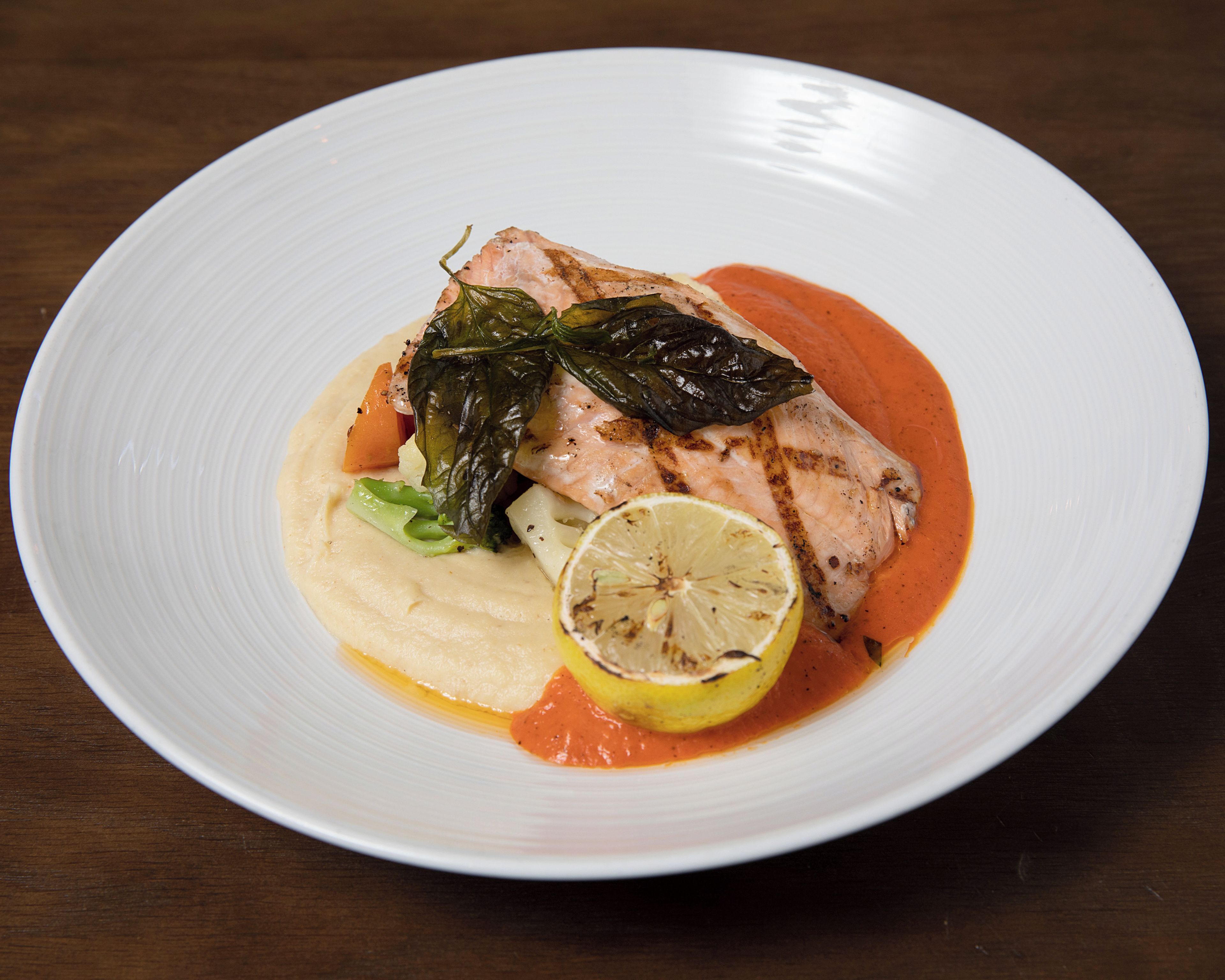 Un plato blanco con un trozo de pescado a la parrilla, una rodaja de limón y salsa cremosa.