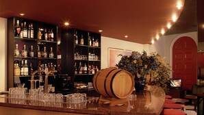 Zona de bar con un barril de madera, varias estanterías llenas de botellas de licores, vasos y un televisor.