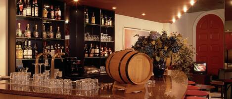 Zona de bar con un barril de madera, varias estanterías llenas de botellas de licores, vasos y un televisor.