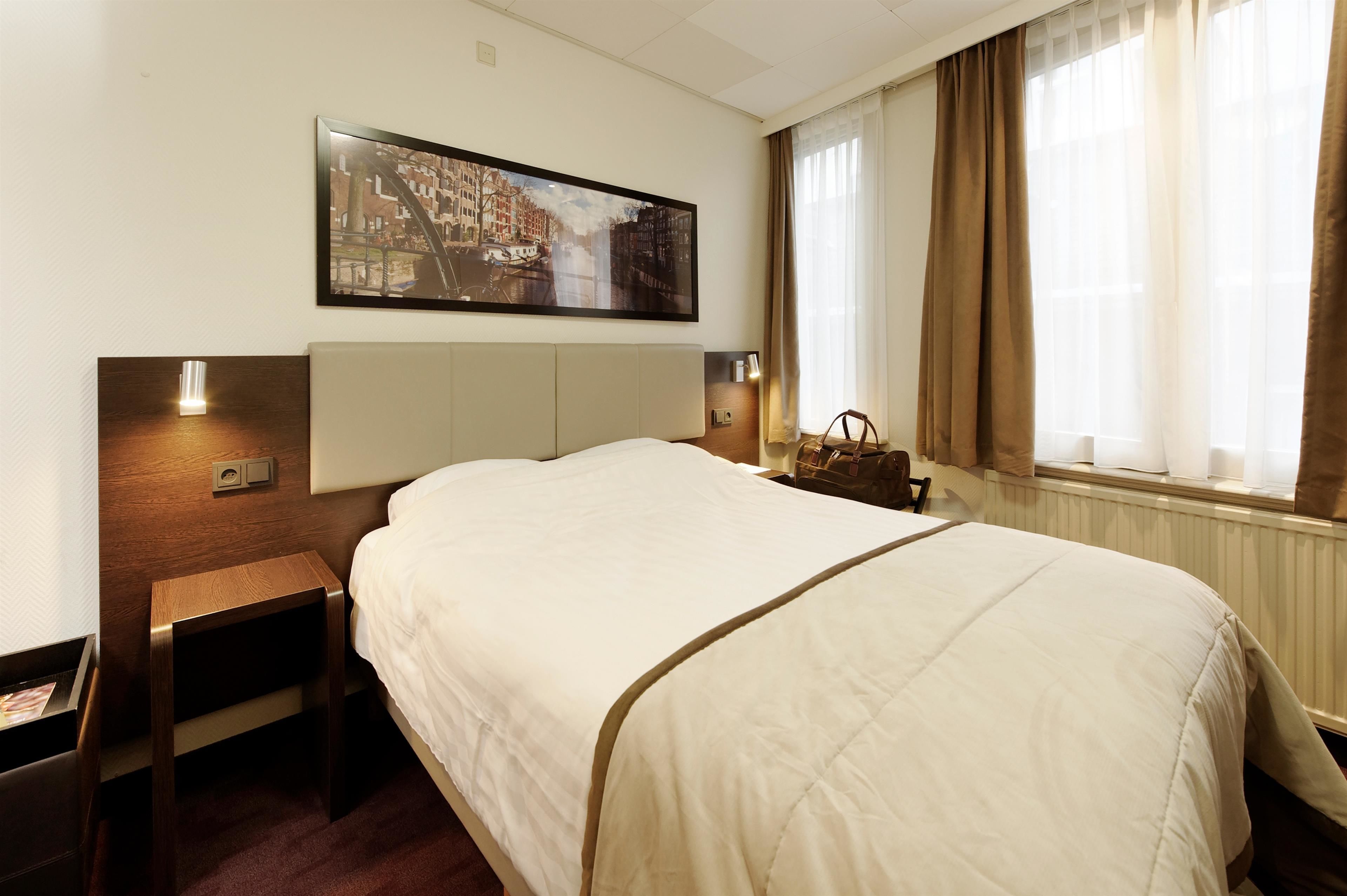 A hotel room with a large bed, a nightstand, a lamp, a painting on the wall, and a window with curtains.