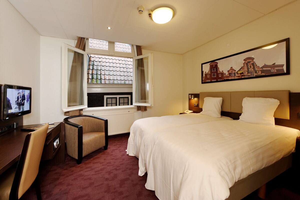 Quarto de hotel com cama, escrivaninha com computador, cadeira, janela com cortinas e um quadro na parede.