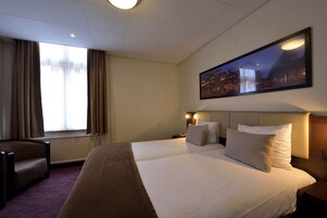Une chambre d’hôtel avec un grand lit, une chaise, une table de chevet, une lampe et une fenêtre avec des rideaux.