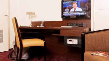 A hotel room with a wooden desk, a television mounted on the wall, a chair, and a small table with a lamp.