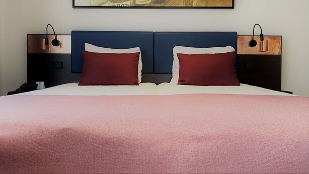 Une chambre d’hôtel avec un grand lit, deux tables de chevet, une lampe fixée au mur et un tableau encadré au-dessus du lit.
