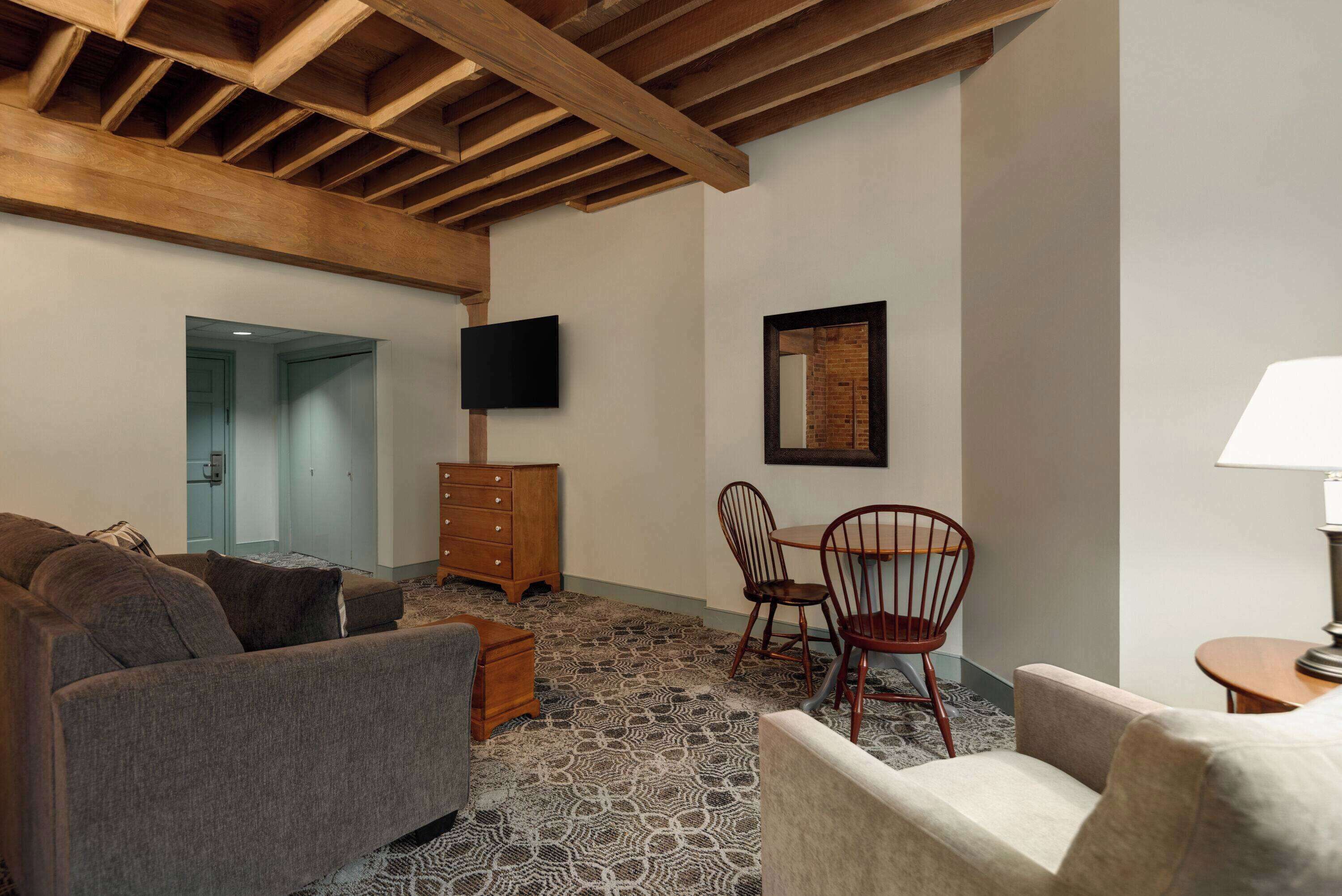 A cozy living room with a patterned carpet, wooden ceiling beams, a flat-screen TV, and a framed picture on the wall.