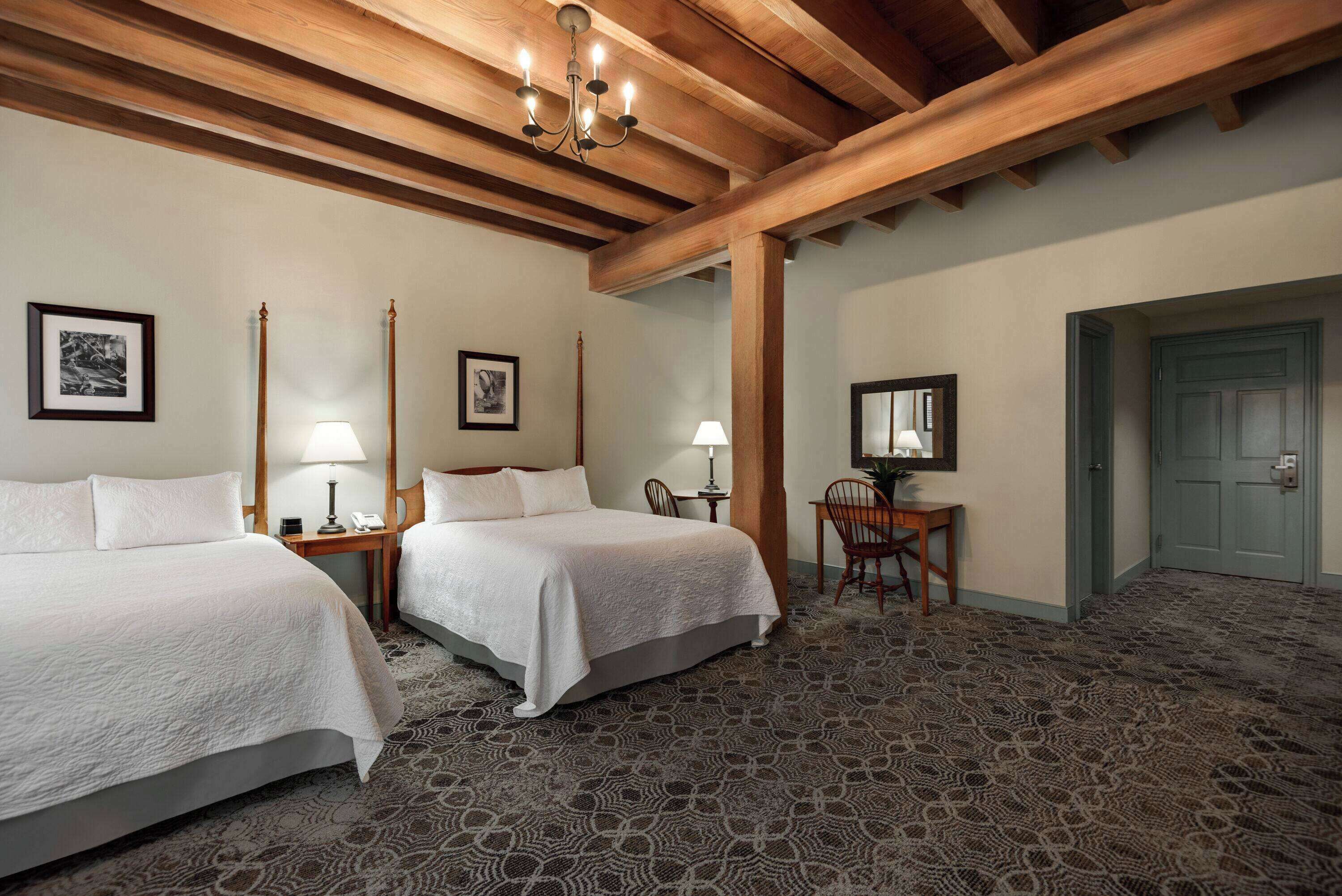 Een hotelkamer met twee bedden, een bureau en een stoel. Er hangen foto's in lijsten aan de muur, een kroonluchter en een deur is zichtbaar op de achtergrond.