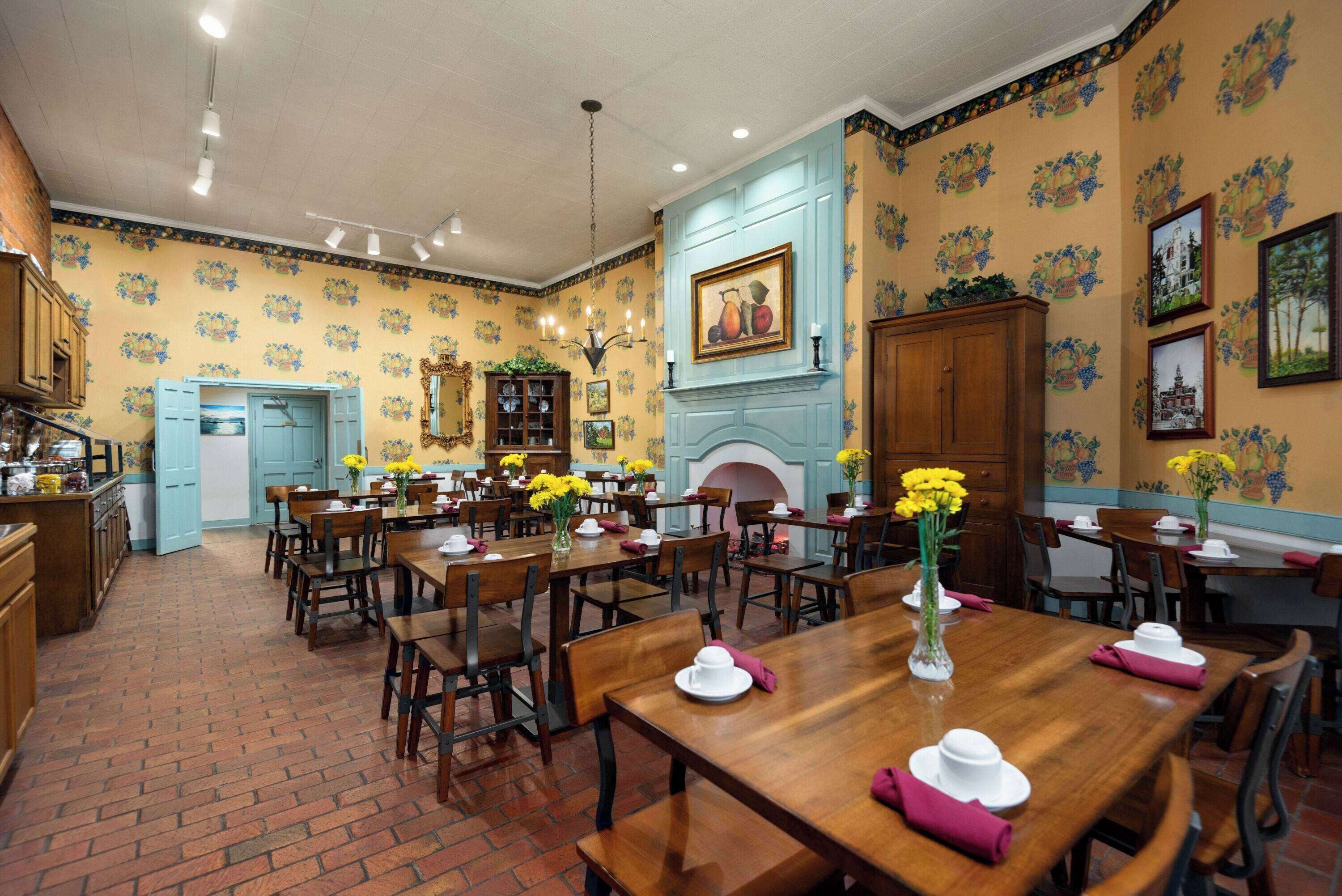Un comedor con suelo de ladrillo, mesas y sillas de madera, y papel tapiz floral amarillo.