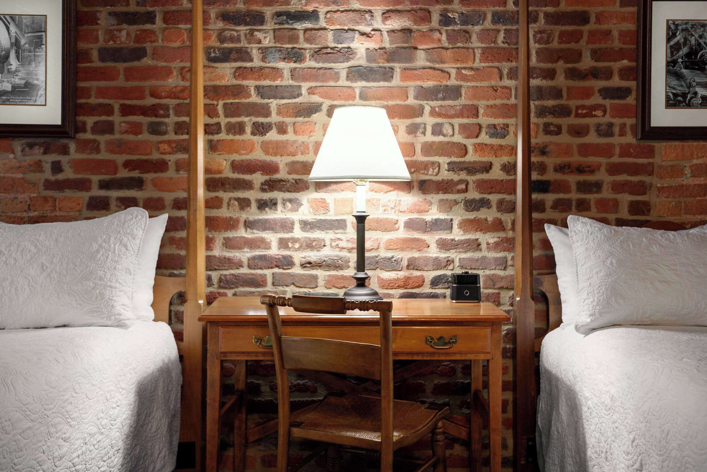 A room with two beds, a wooden desk, a lamp, and a brick wall.
