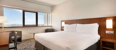 A hotel room with a large bed, a desk with a chair, a small table, and a nightstand. There is a window with a view of the sky and distant landscape.