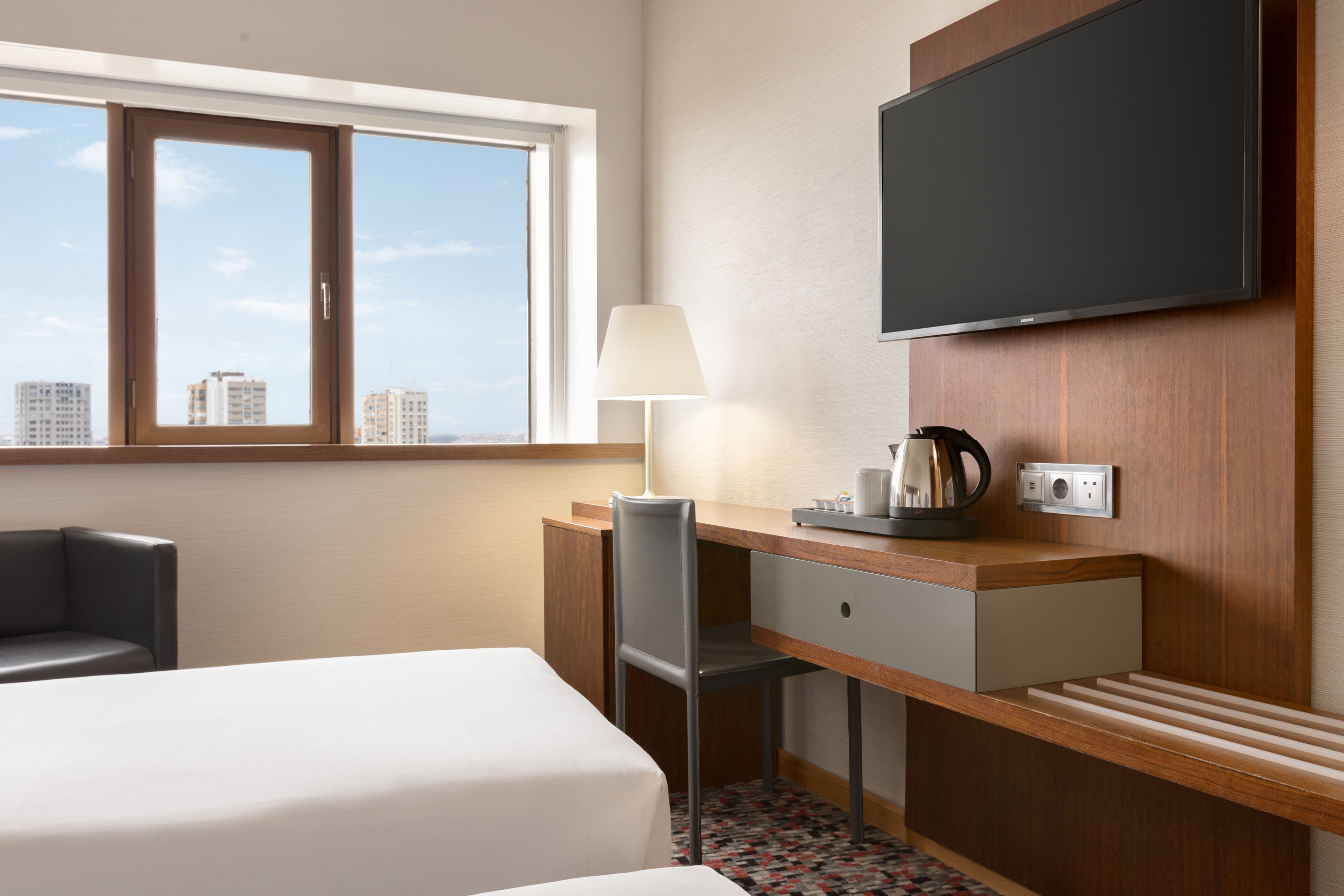 A hotel room with a bed, desk, chair, lamp, and a flat-screen TV mounted on the wall.