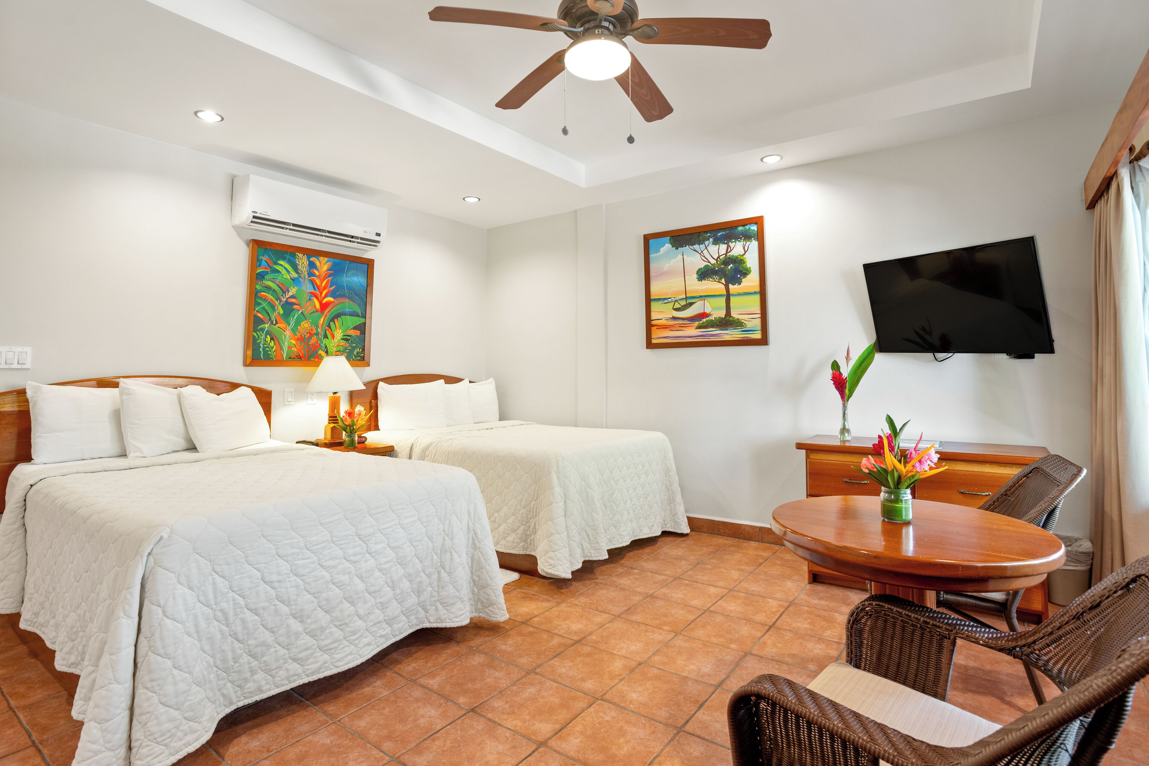 A hotel room with two beds, a ceiling fan, a TV, a dining table with chairs, and artwork on the walls.