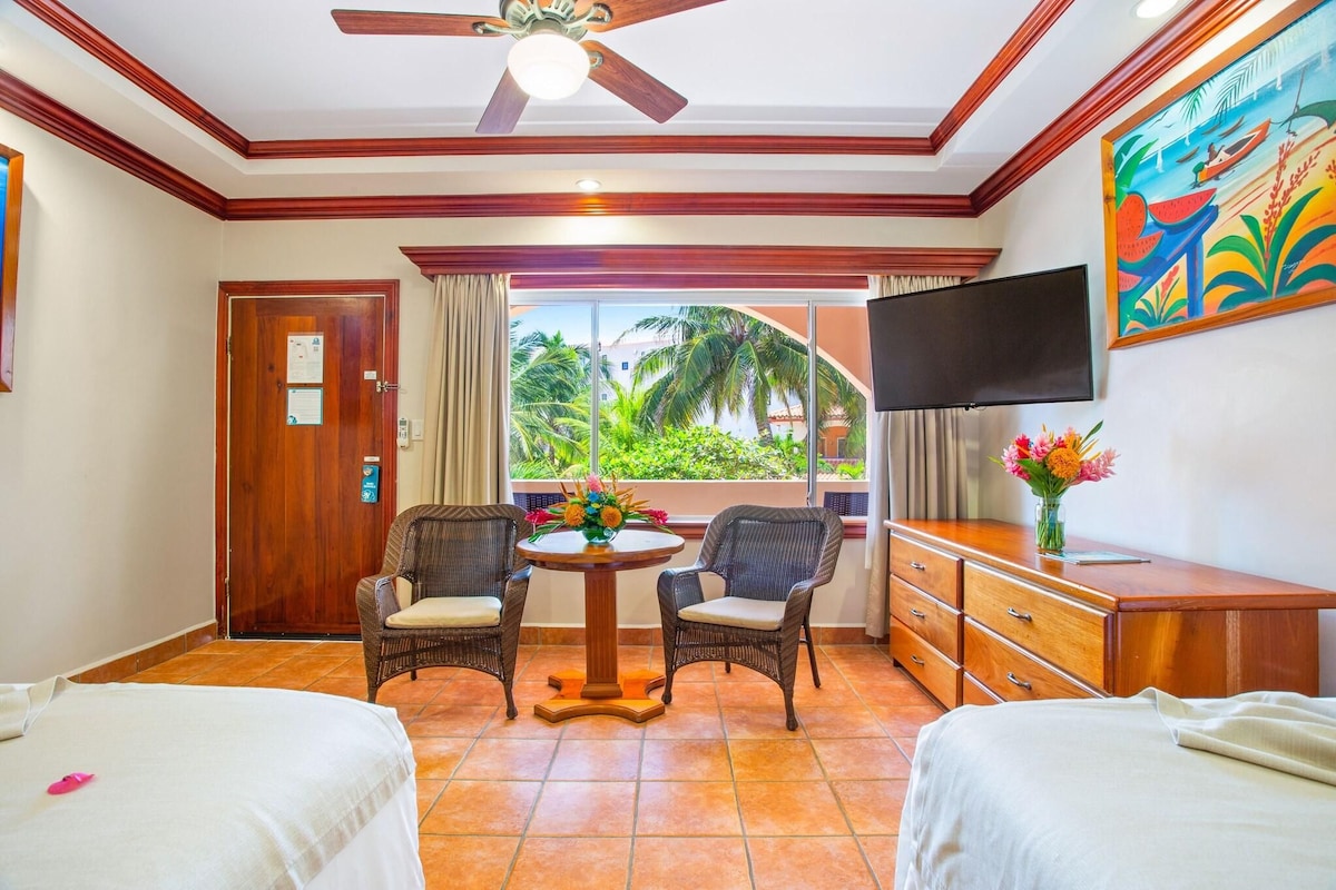 A hotel room with two beds, a dining table with chairs, a TV, and a view of palm trees.