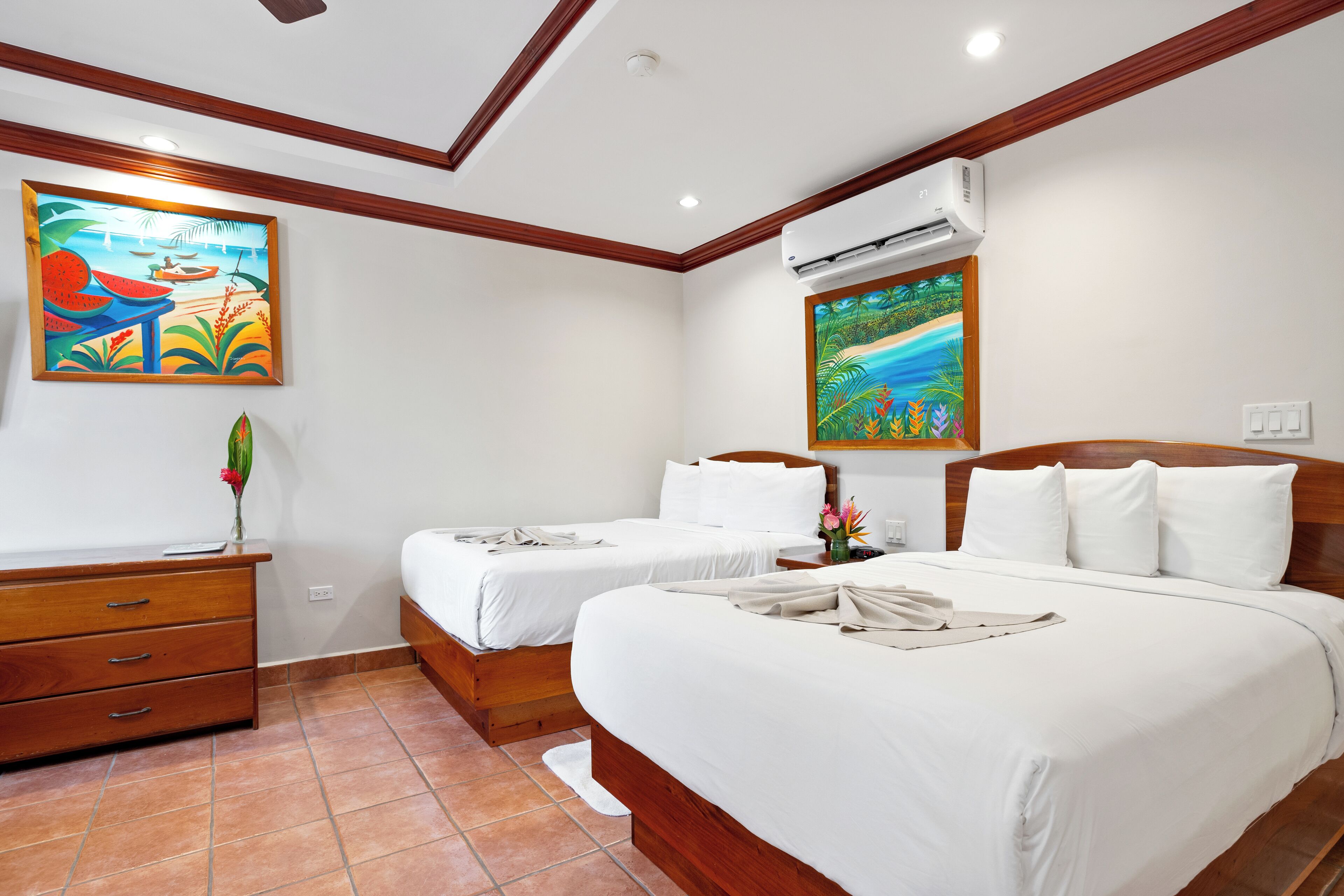 A hotel room with two beds, wooden headboards, white bedding, a wooden dresser, and two paintings on the walls.