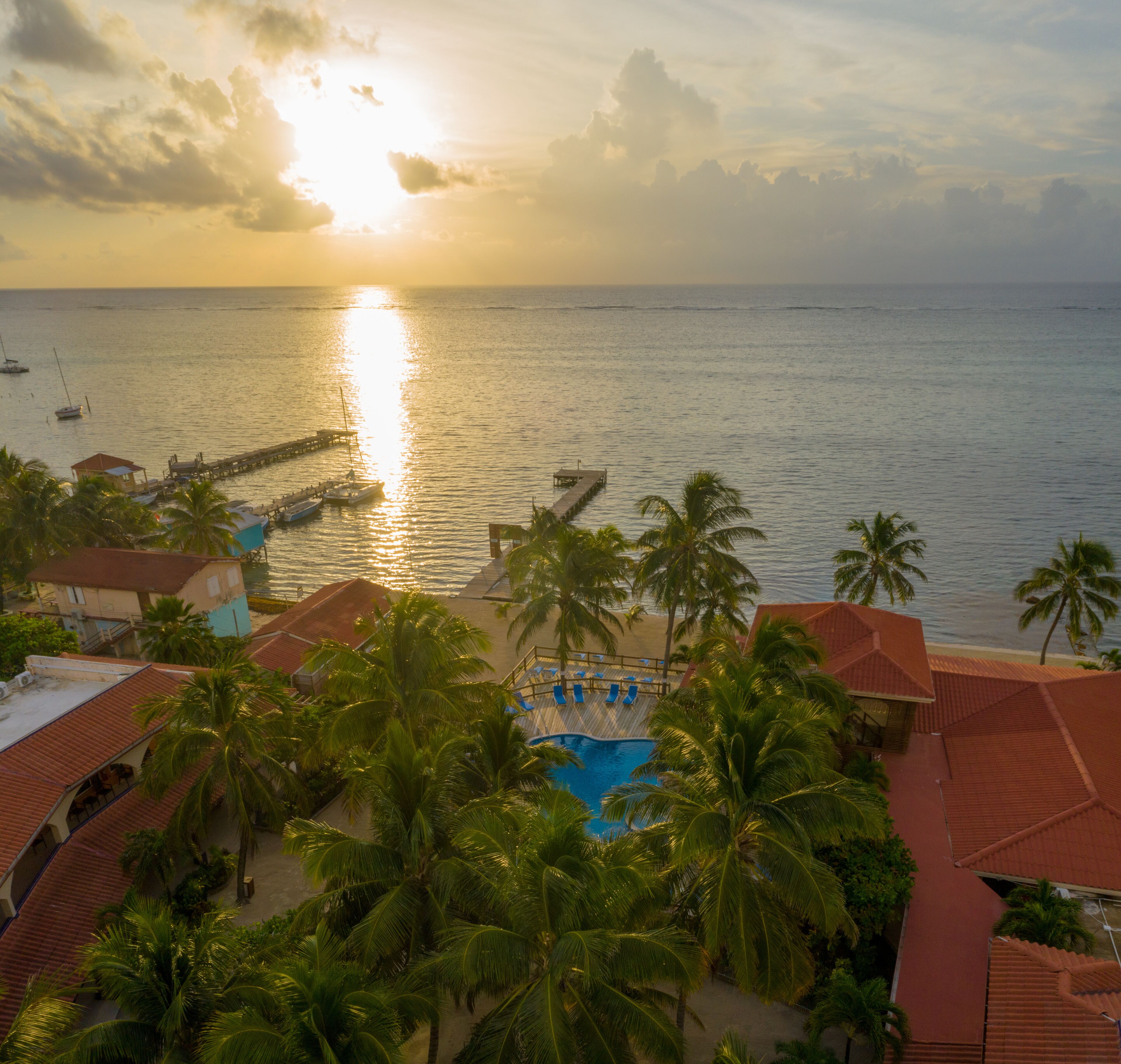A resort with a swimming pool, palm trees, and a view of the ocean at sunset.