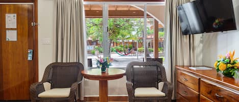 A room with two wicker chairs, a wooden table, a TV mounted on the wall, and a view of an outdoor area with a hammock and greenery.