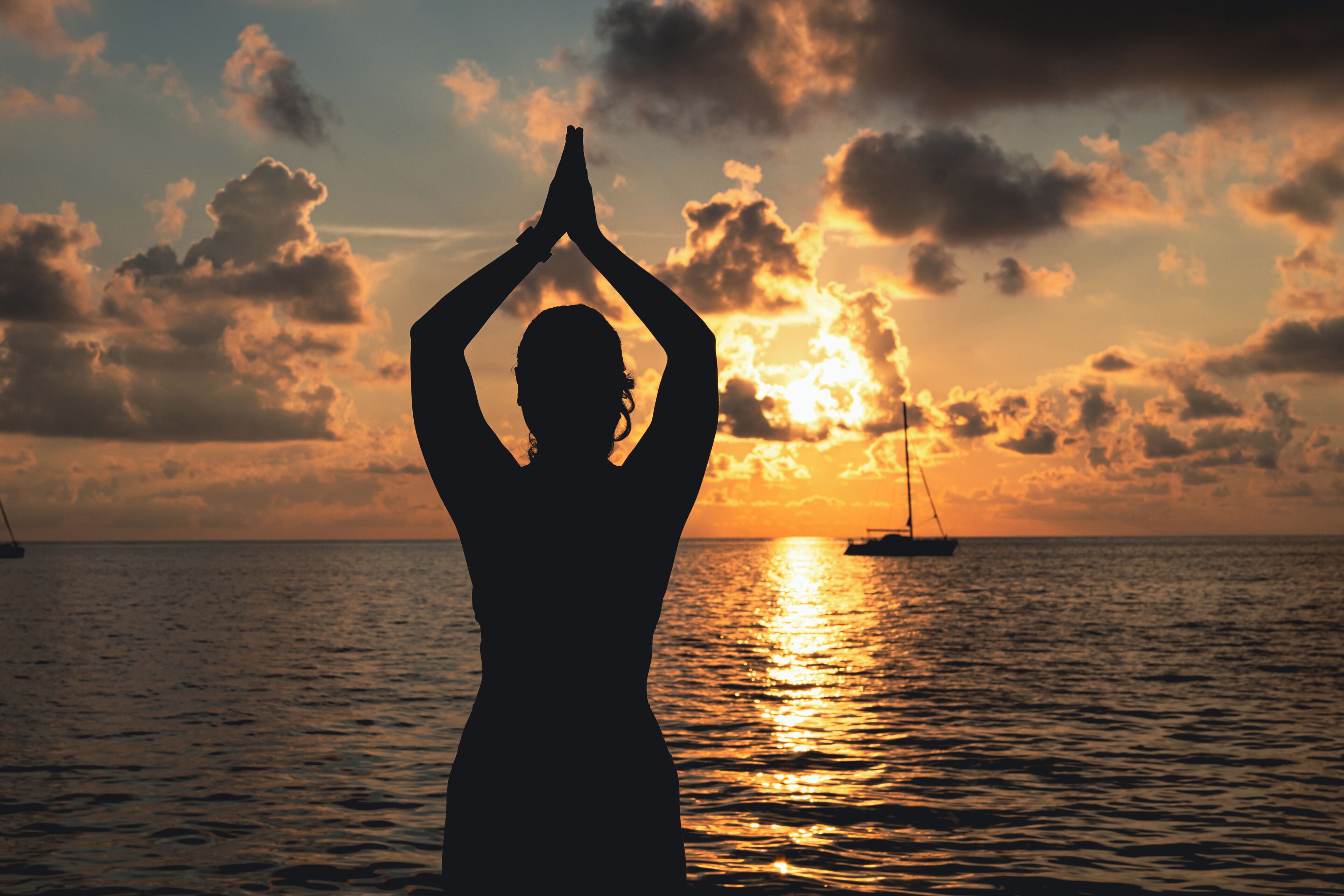 La silhouette d’une personne pratiquant une posture de yoga, se détachant sur le ciel orangé d’un coucher de soleil sur l’océan, avec un voilier visible au loin.