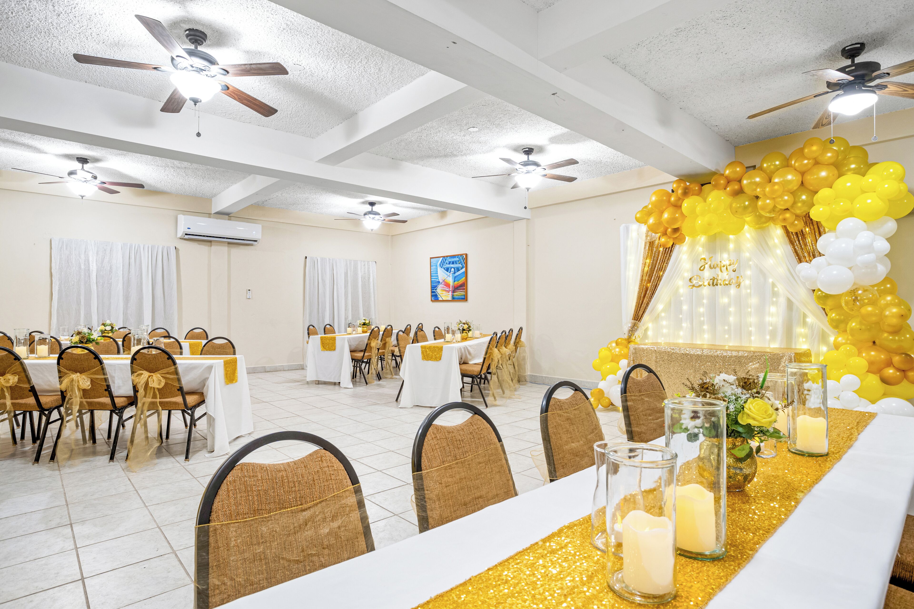 A banquet hall with white tables, gold chairs, ceiling fans, and a yellow and white balloon arch.