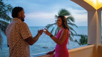 A couple toasting with drinks on a balcony overlooking the ocean with palm trees in the background.