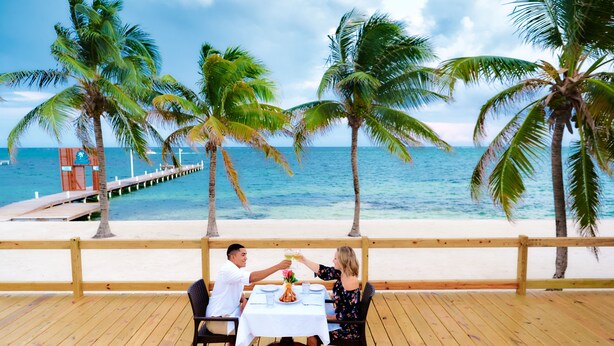 A wooden deck with a table set for two, overlooking a beach with palm trees and the ocean. - Sunbreeze Hotel (San Pedro)