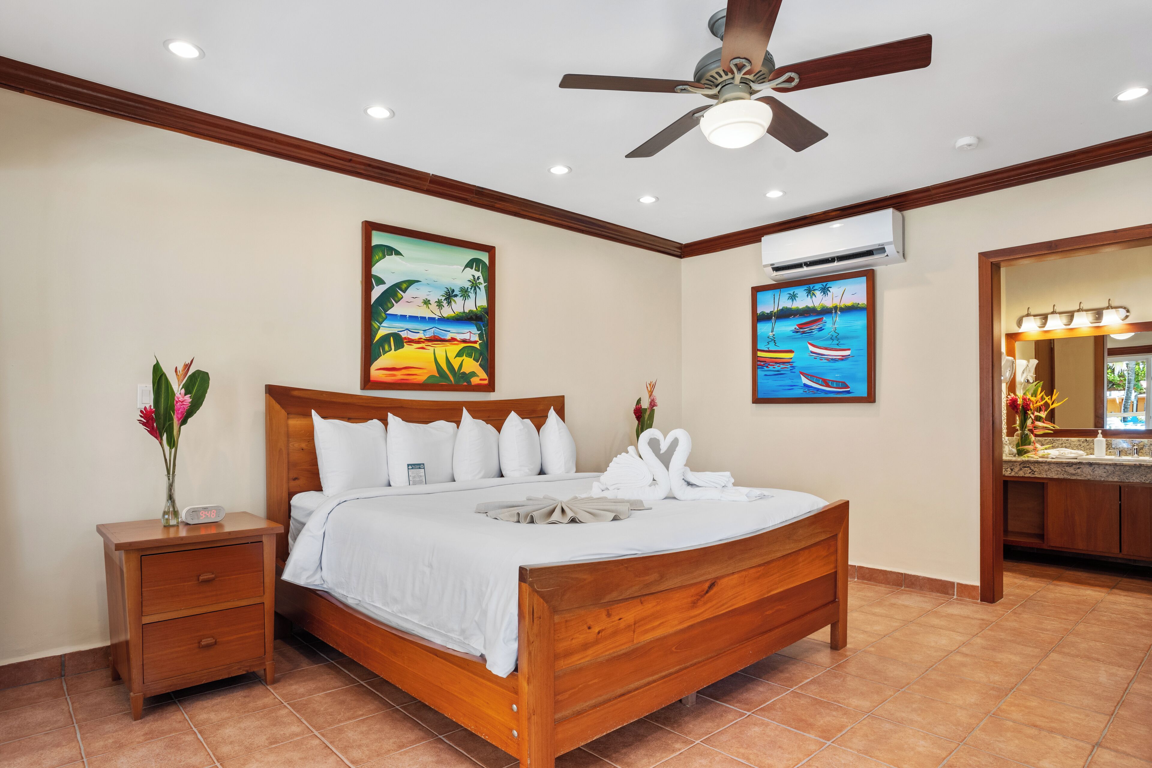 A wooden bed with white bedding, a nightstand with a vase of flowers, and two paintings on the wall depicting tropical scenes.