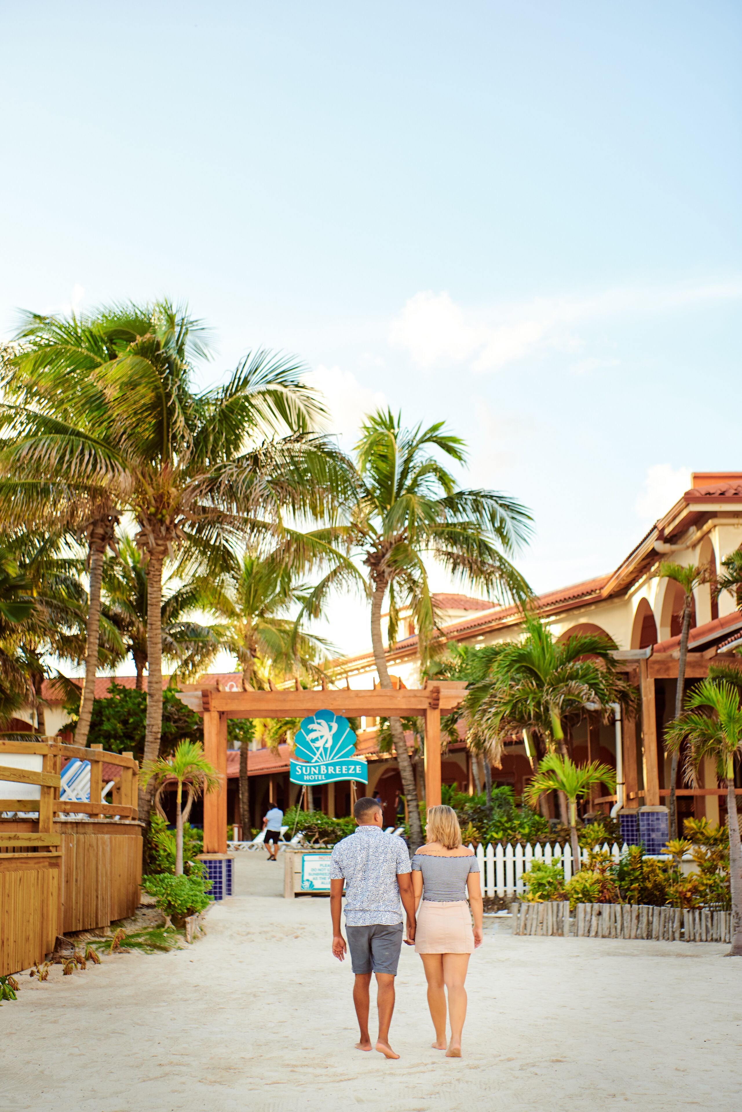 A couple walking towards a resort entrance with palm trees and a white picket fence.