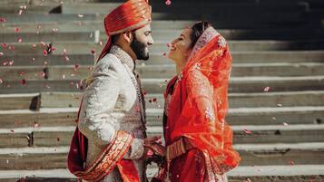 Un couple, vêtu de tenues traditionnelles de mariage, se tient sur des marches, tandis que des pétales de fleurs tombent autour d’eux.