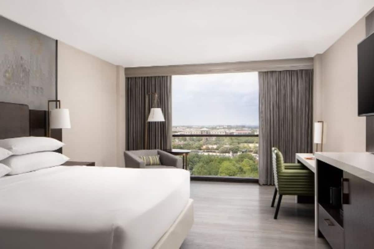 A hotel room with a large bed, a desk, a chair, and a TV. There is a window with a view of a cityscape.