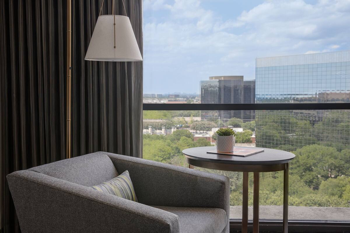 A modern room with a grey sofa, a small round table with a plant, and a view of a cityscape through a large window.