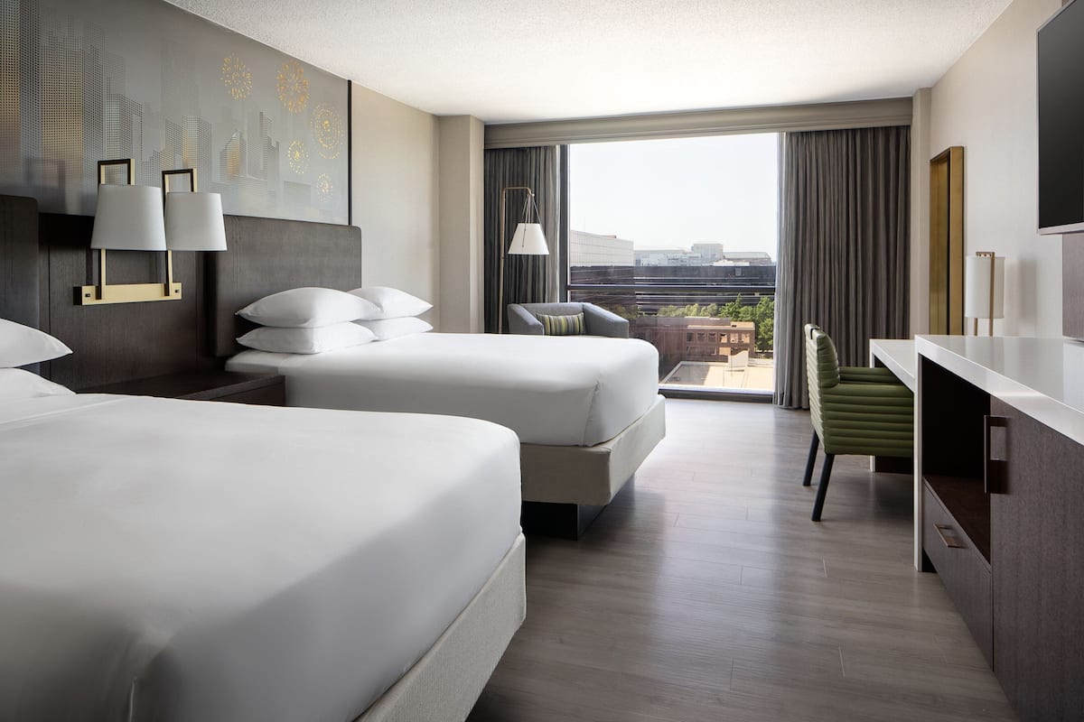 A hotel room with two beds, a desk, a chair, and a large window with a city view.