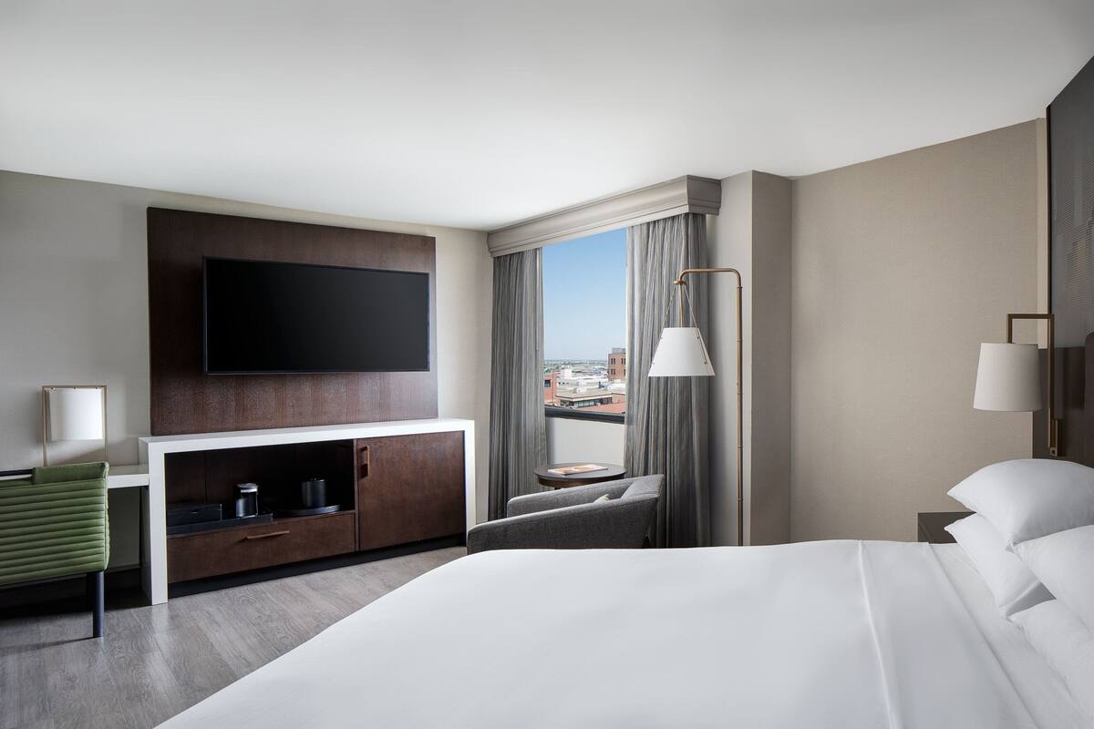 A hotel room with a large bed, a flat-screen TV mounted on a wooden panel, a small table with a lamp, a green chair, and a view of the city through large windows.