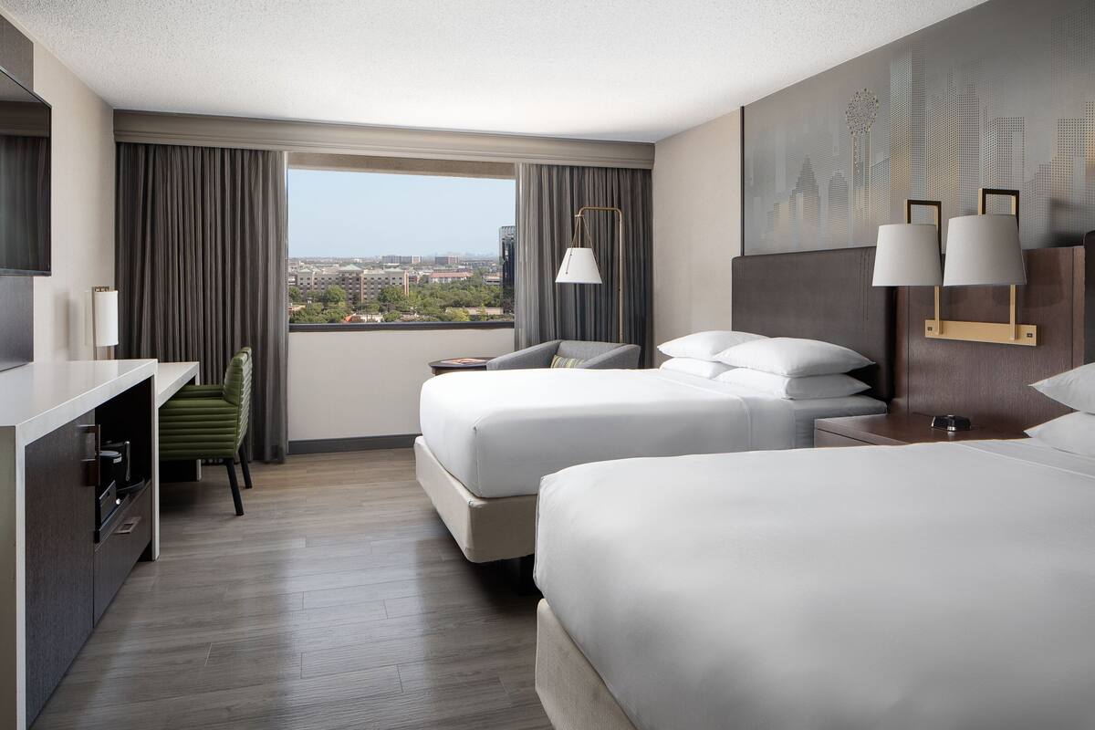 A hotel room with two beds, a desk, a chair, and a large window with a city view.