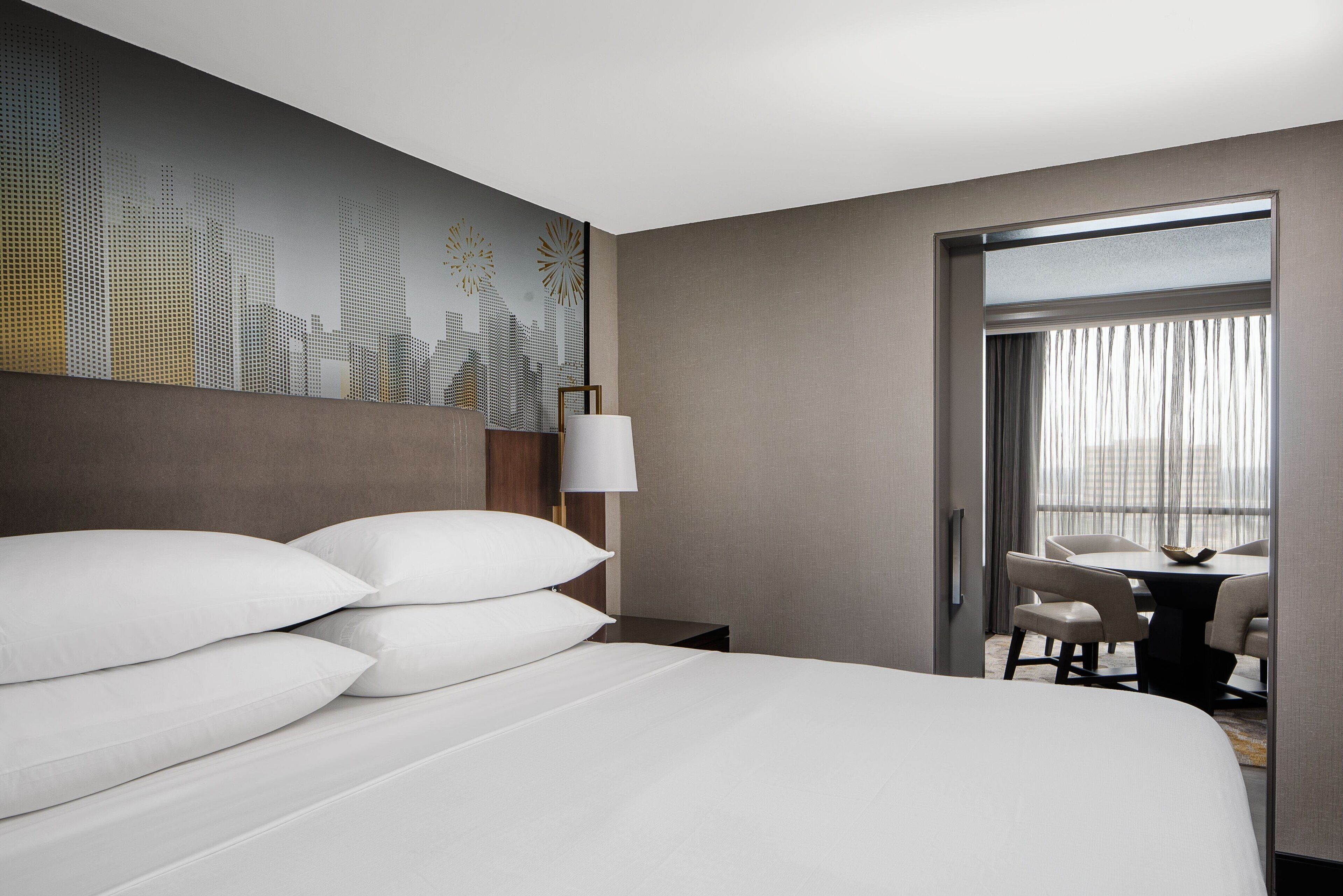 Habitación de hotel con una cama grande, una mesita de noche, una lámpara, un área de comedor con sillas y vistas a través de una puerta.