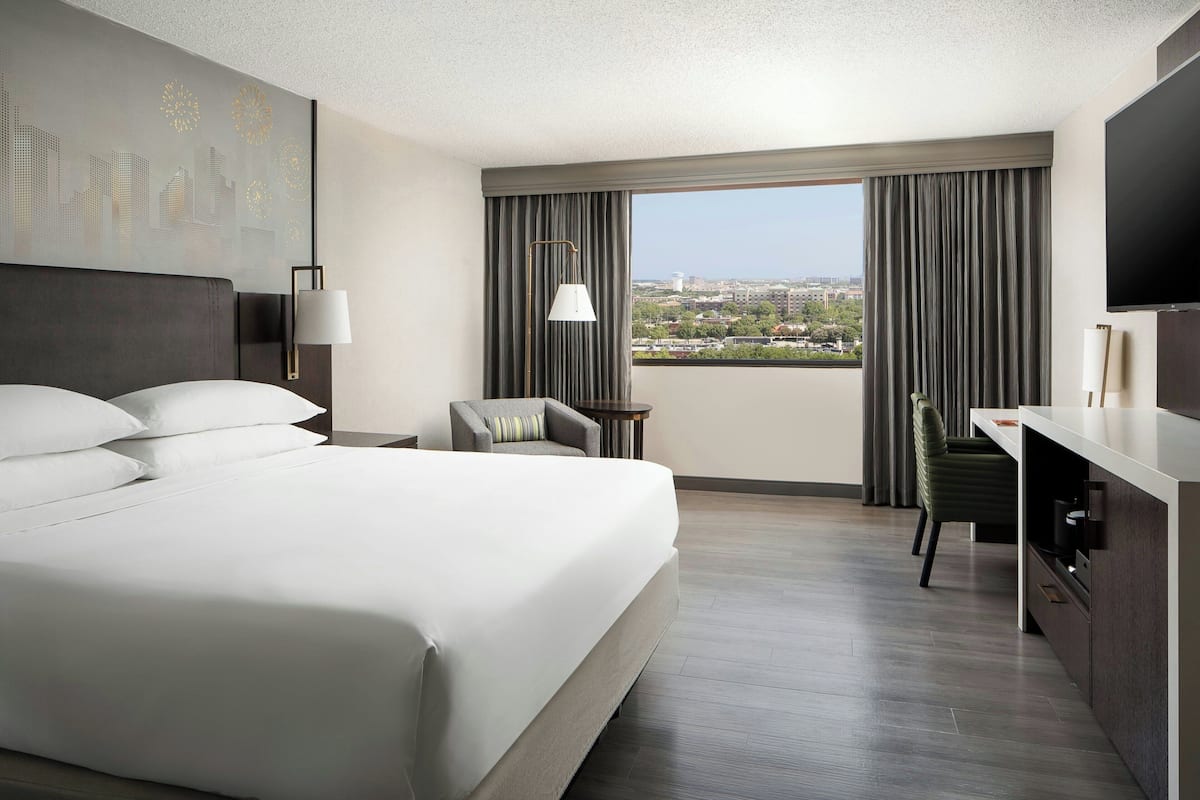 A hotel room with a large bed, a desk, a chair, a TV, and a view of the city.