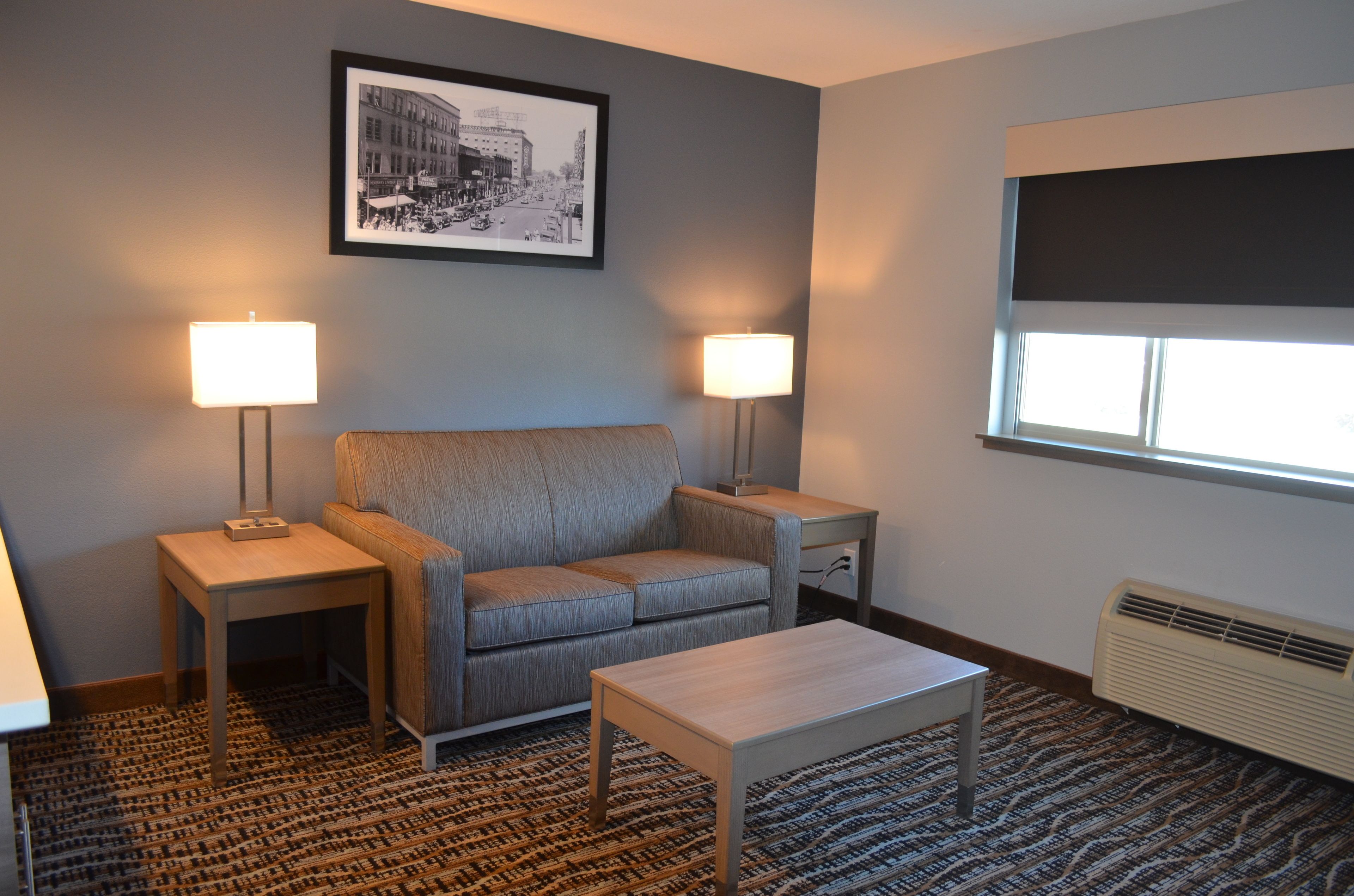 A hotel room with a grey sofa, two bedside tables with lamps, a small wooden table, a patterned carpet, a framed black and white cityscape picture on the wall, and a window with a dark blind.