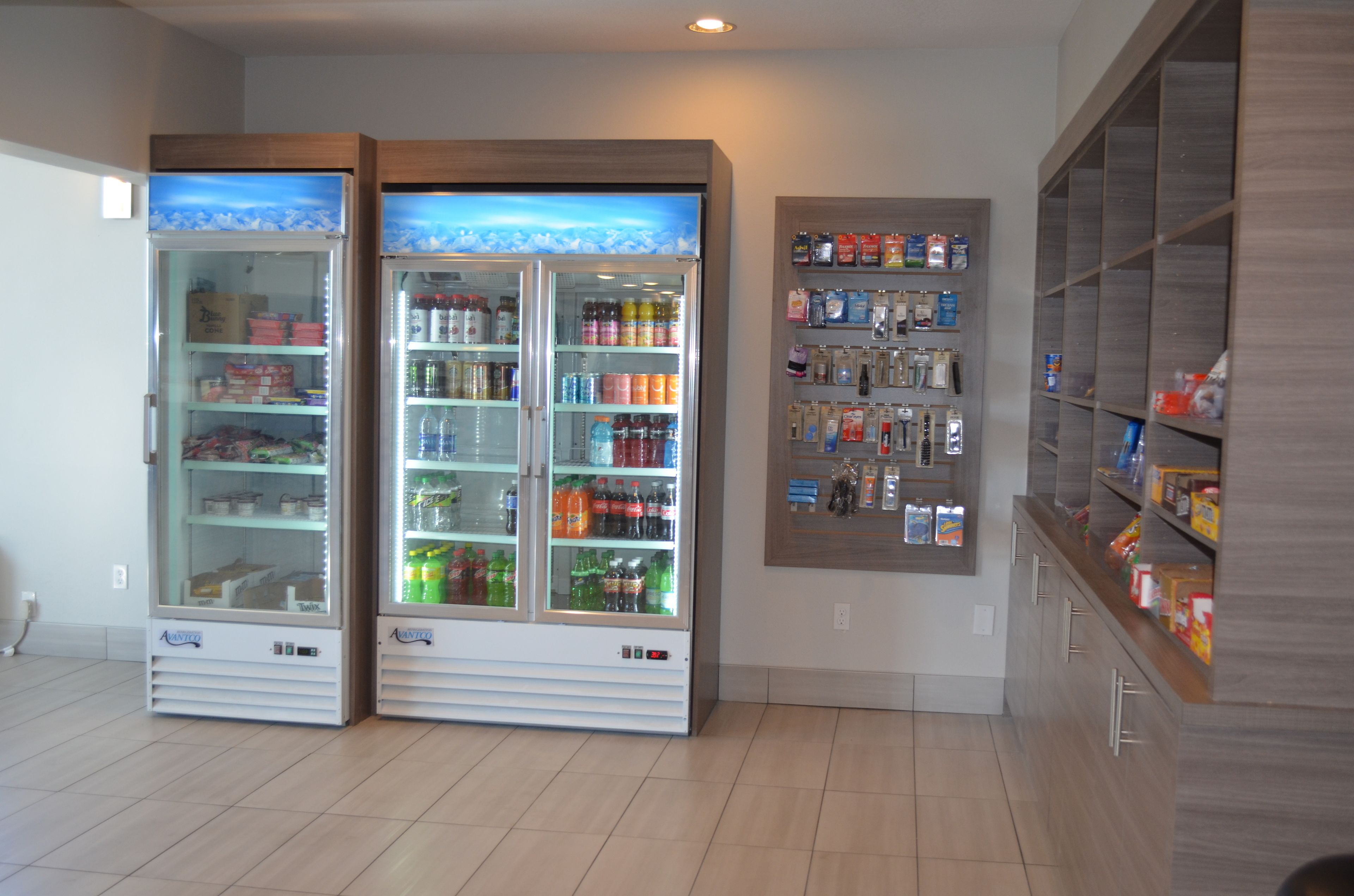 Two beverage vending machines with a variety of drinks and snacks, a wooden cabinet with more snacks, and a tiled floor.
