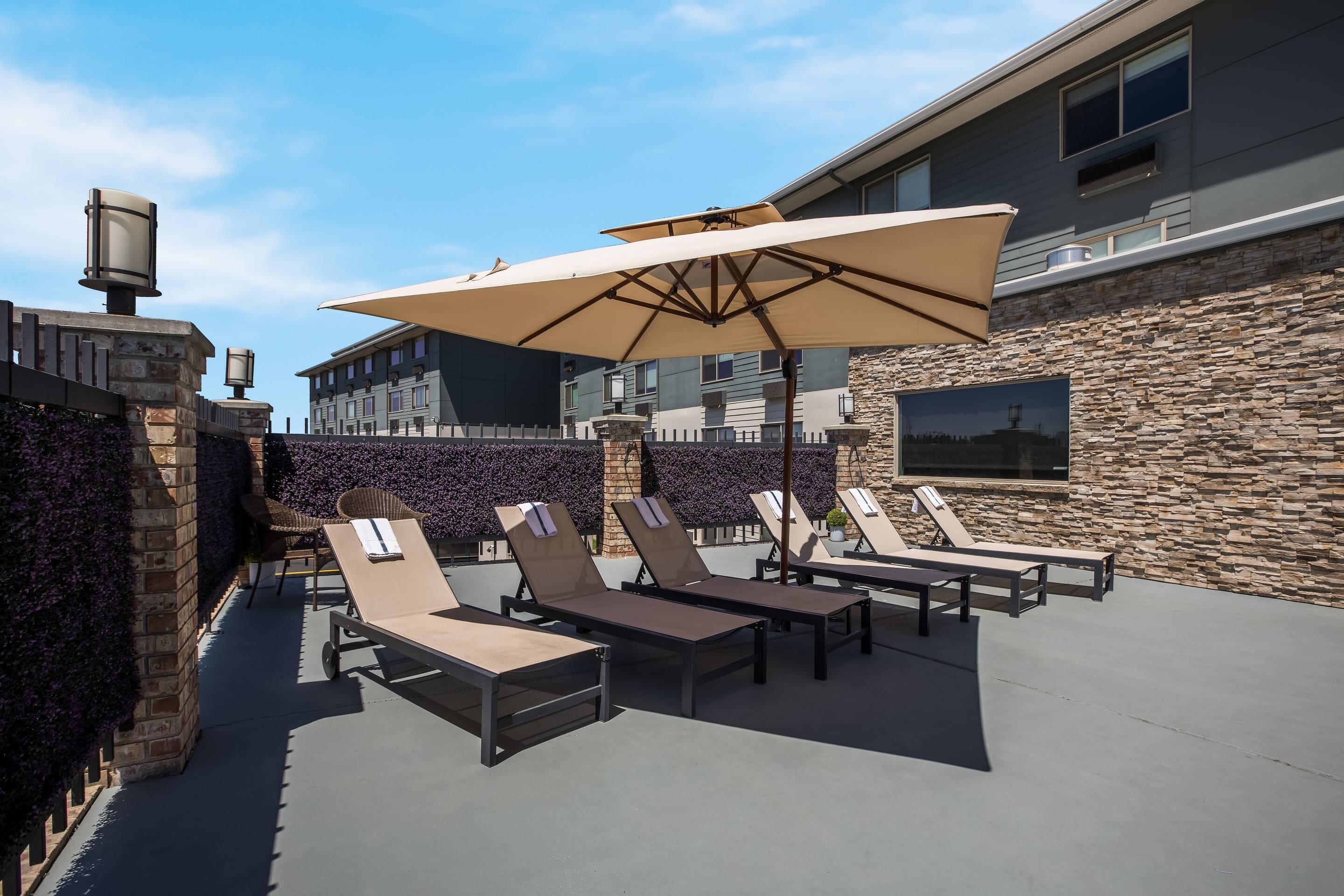 An outdoor patio with lounge chairs and a large umbrella, stone walls, and a modern building in the background.