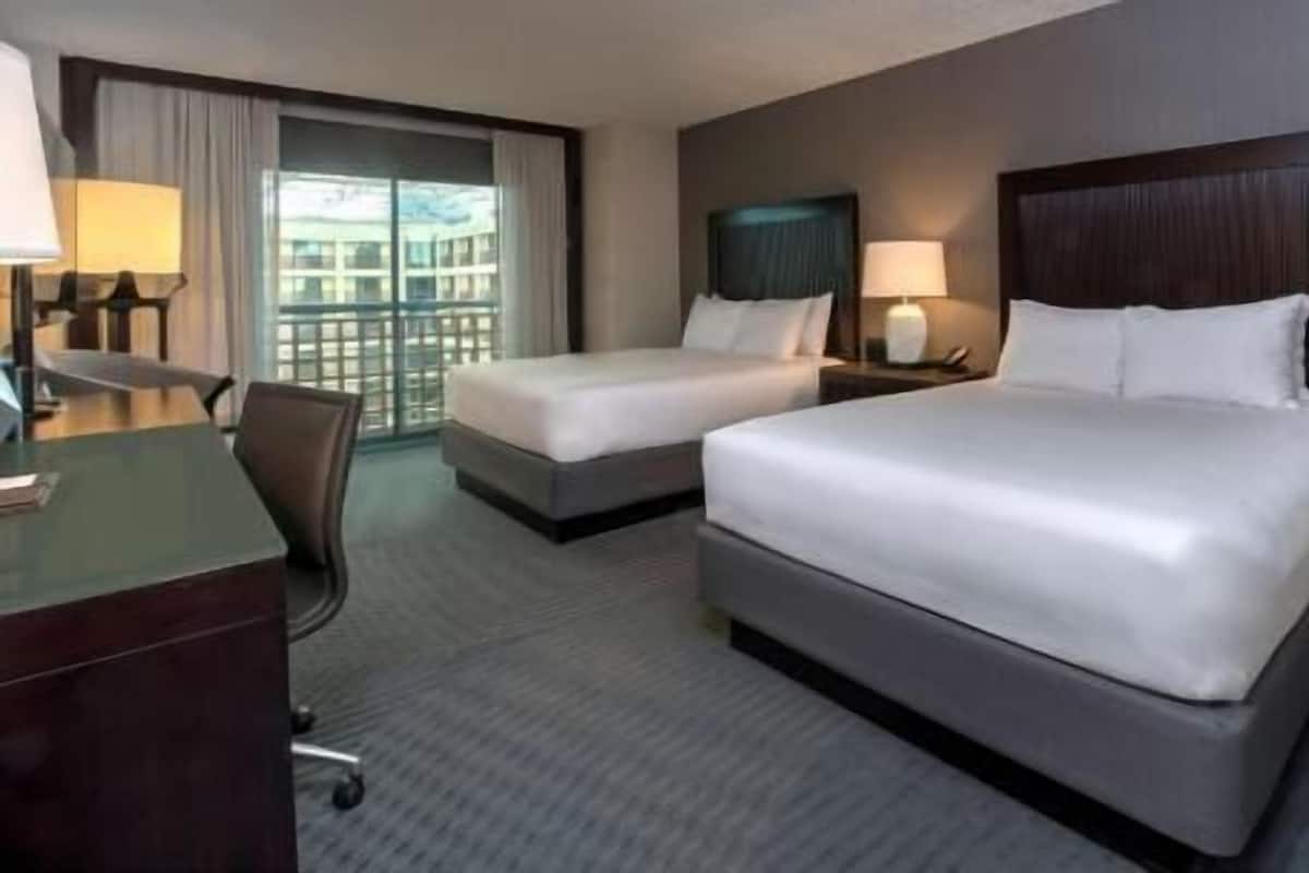 A hotel room with two beds, a desk, a chair, a window with curtains, and a view of buildings outside.