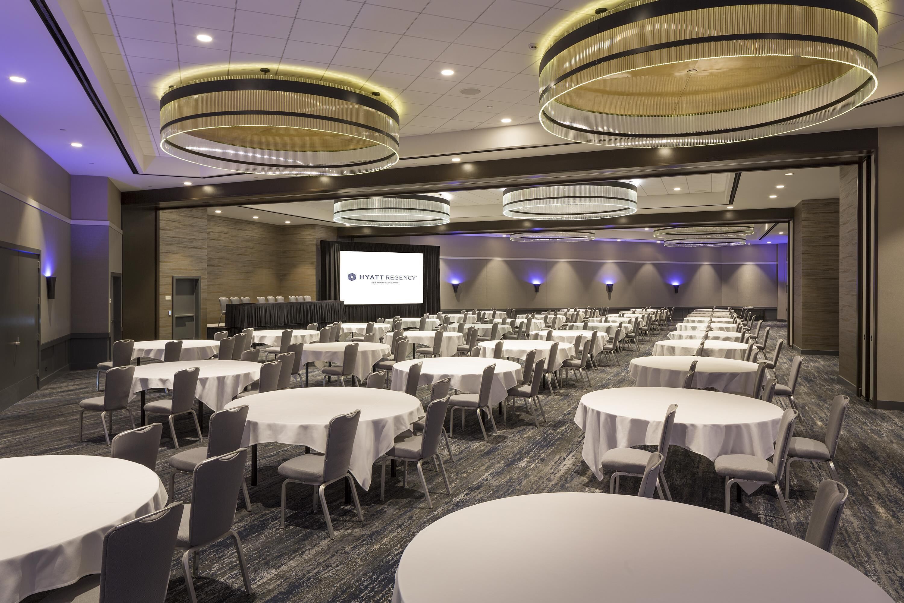 Un salón de conferencias con varias mesas redondas y sillas, una pantalla grande que muestra 'HYATT REGENCY', y luminarias modernas.